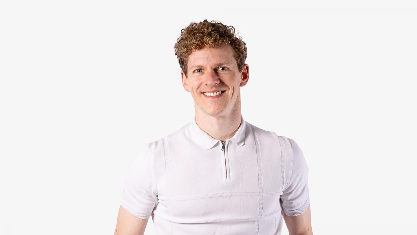 Man with fair curly hair, wearing a white short-sleeved shirt, looks directly at the camera and smiles warmly and open mouthed