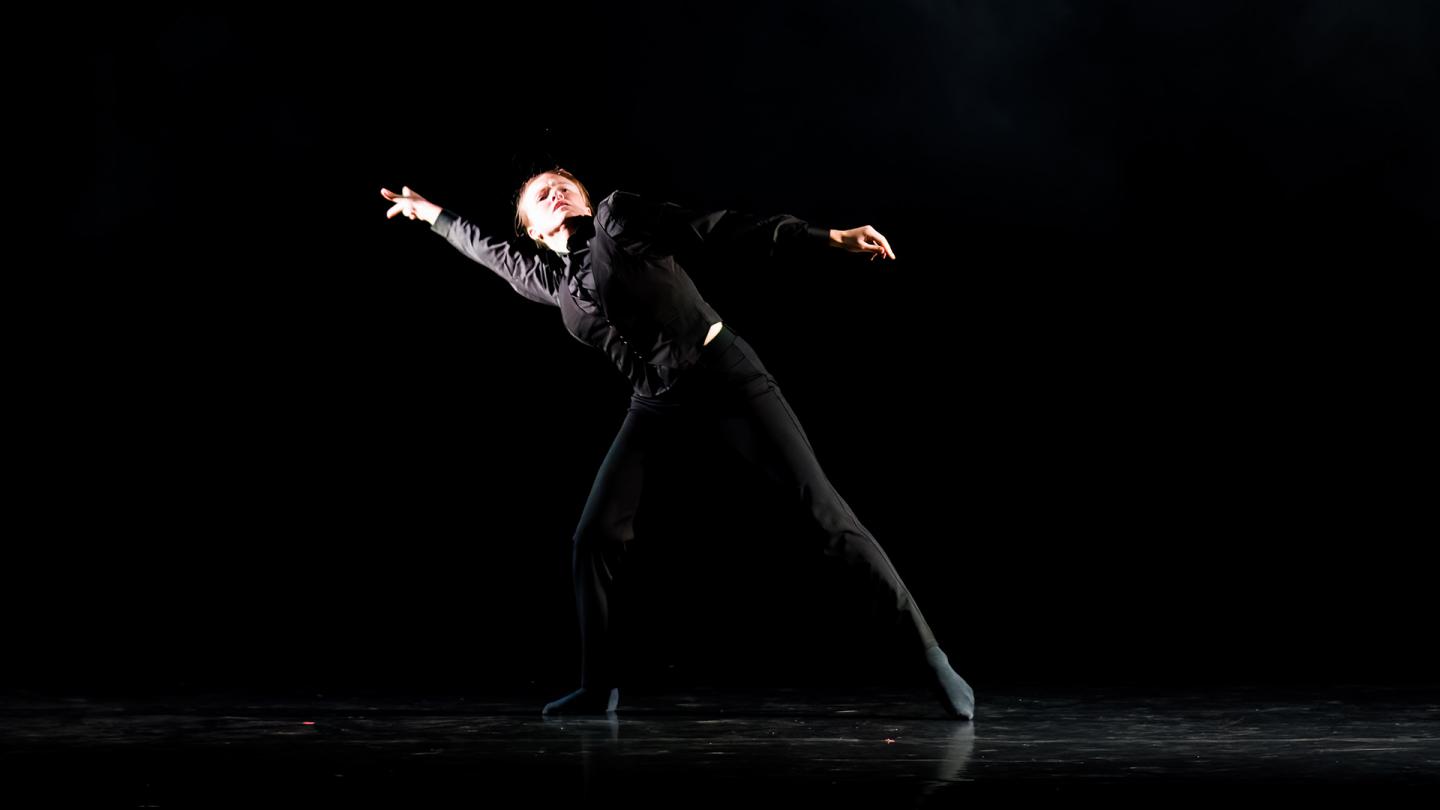 Dancer in a dark top and trousers, against a darkened stage, stands with legs apart, leaning to their right with an arm outstretched