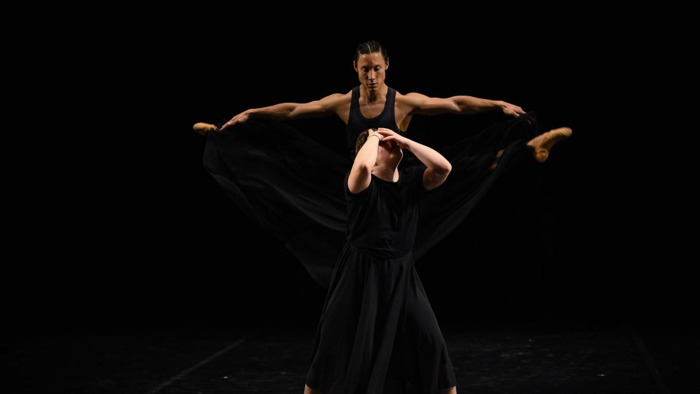 A dancer holds his face as another dancer leaps behind him with outstretched legs, looking down on the other dancer.