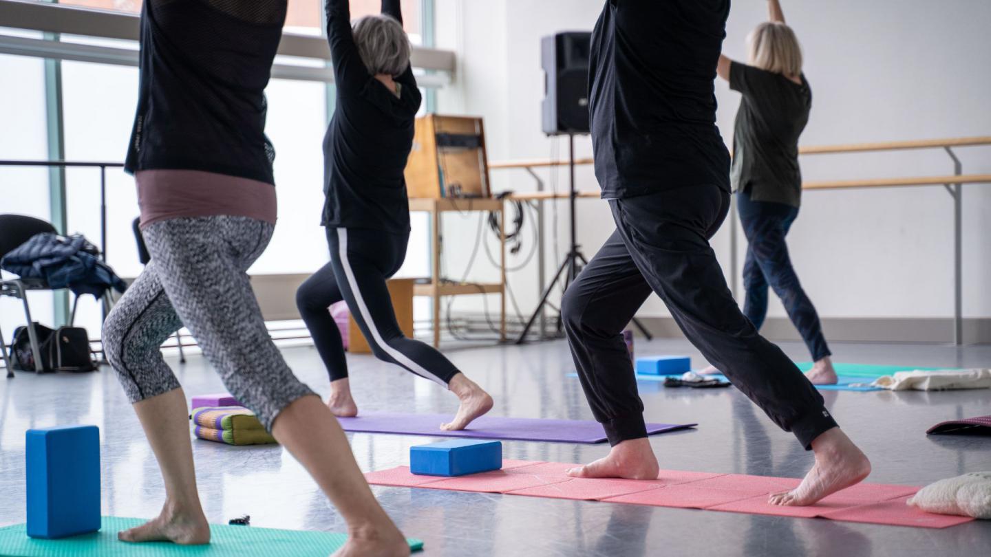 A group of people on yoga mats in a ballet studio. They lift their arms above their head and extend one leg back whilst bending the other.