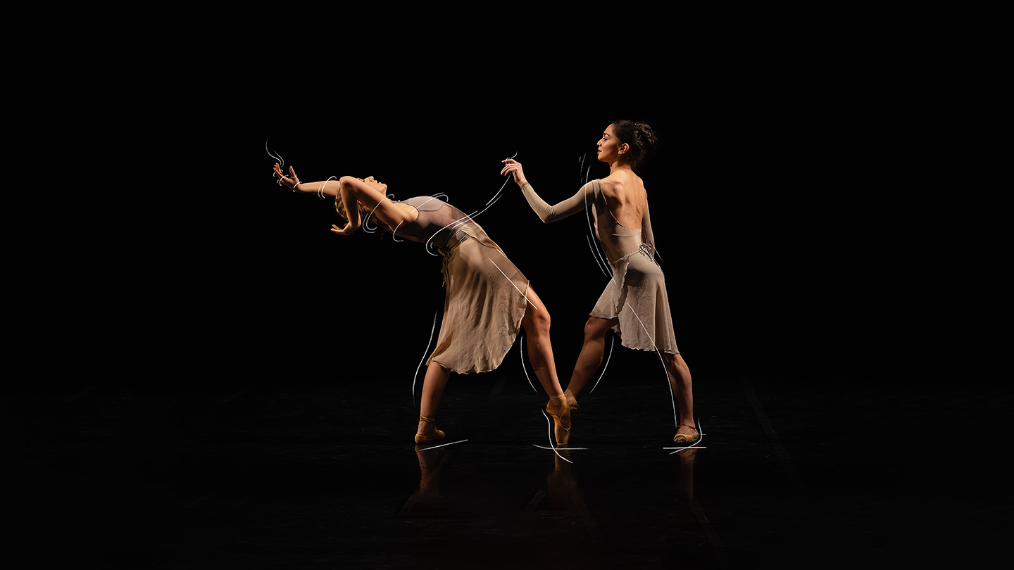 Against a black backdrop two dancers stand facing each other with one foot en pointe, and one dancer leaning back with her arms above her head
