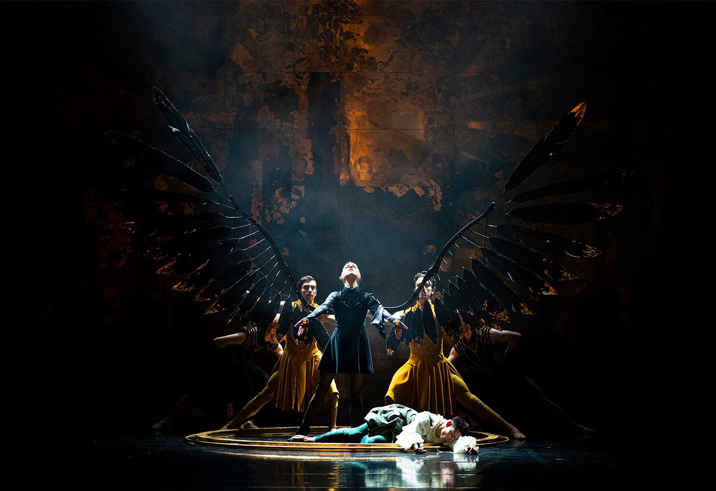 A group of dancers on stage. One is wearing wings and looking up into the sky.