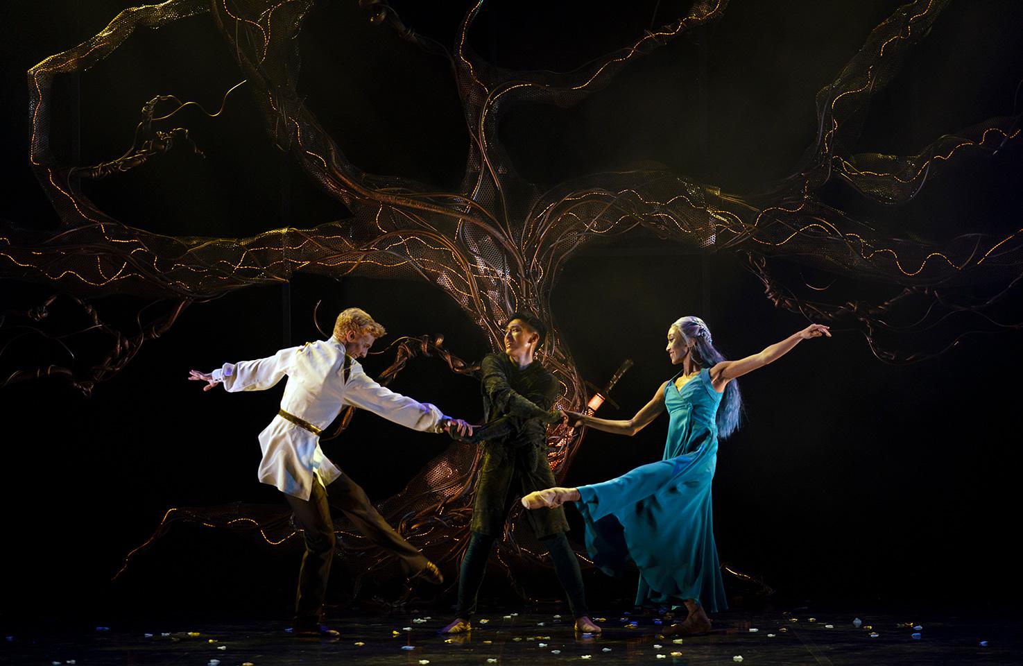 Three dancers perform in front of a large lit tree. Two dance around the other stood in the centre, holding their hands.