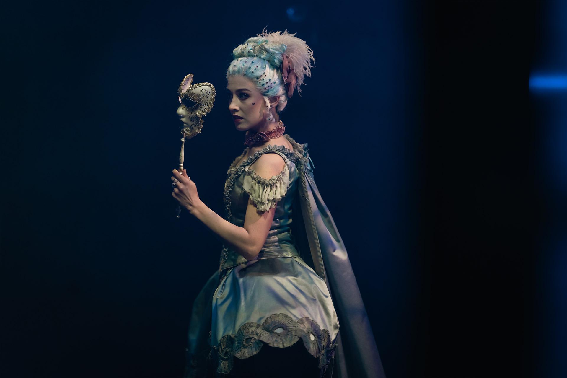 Madame de Pompadour dressed in powder-blue silk removing her ornate mask for a clearer look at something out of frame