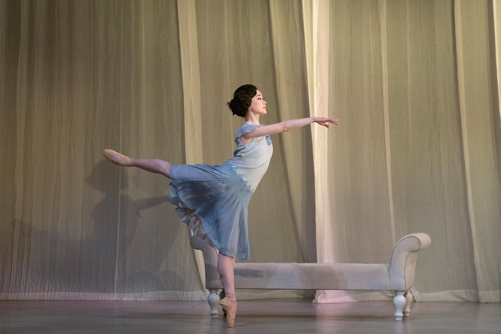 Daisy holding on to the chaise longue while in arabesque and en pointe wearing a powder blue dress