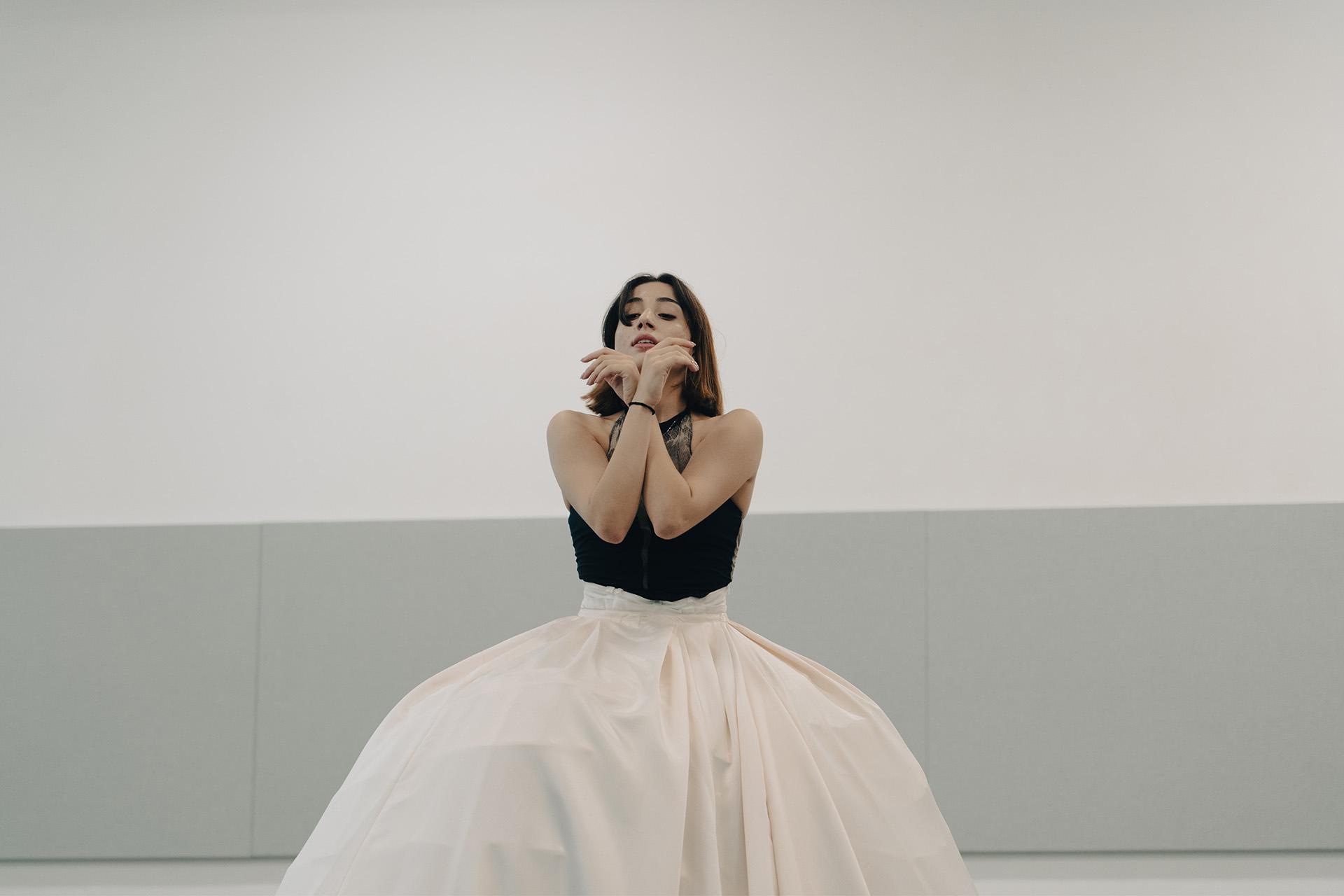 Dancer wearing a voluminous white skirt holding her forearms crossed in front of her face.