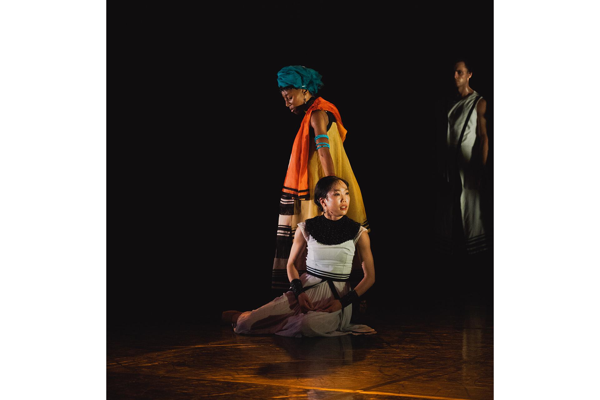 Dancer kneels on the floor while another stands behind her