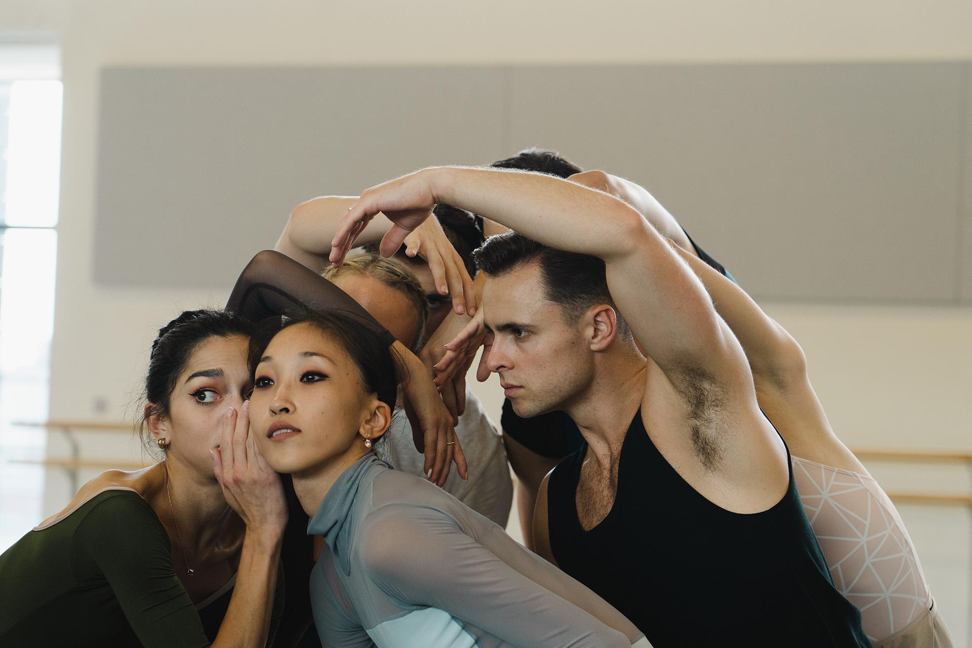 Dancers leaning close to one another, one whispering into another's ear while the rest watch