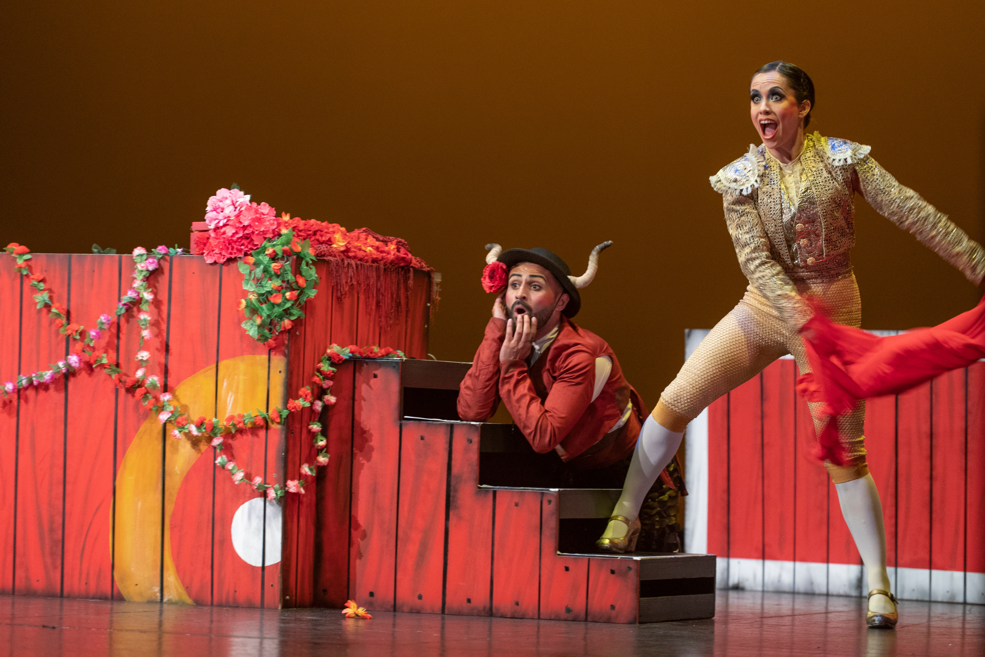 Two dancers on a stage, surrounded by red pieces of set including a small stair case. One dancer is nelt down behind the stair case wearing a red jacket and bull ears on their hat. They look shocked. The other dancer is stood up with their right foot on the staircase, wearing a gold outfit, looking shocked and they have pulled away a red cape.