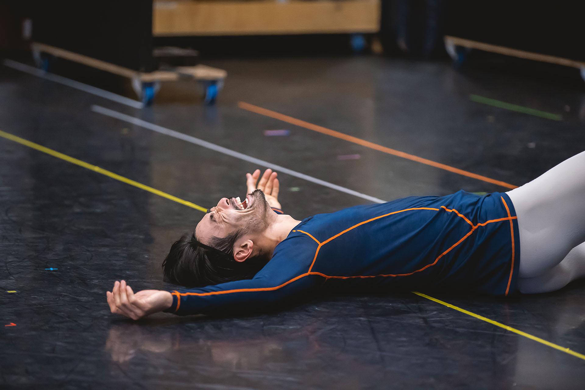 Male dancer wearing a blue top collapsed on his back on black dance floor laughing