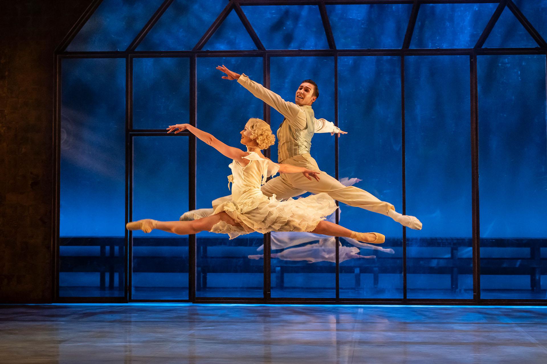 A male and female dancer both performing grand jetés joyously while wearing while clothes against a blue background