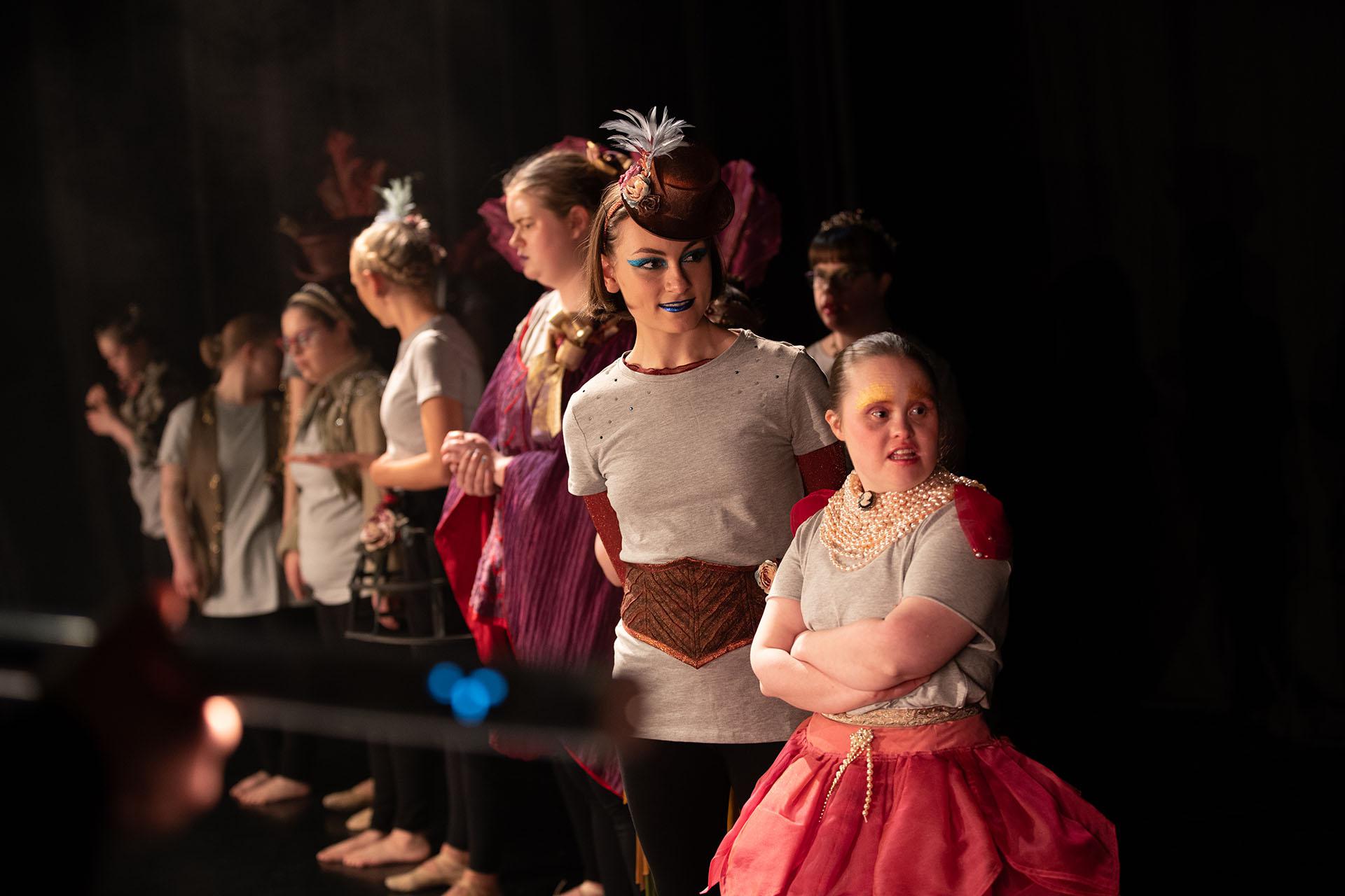 Performers prepare for more filming, all stood in a row against a black background.