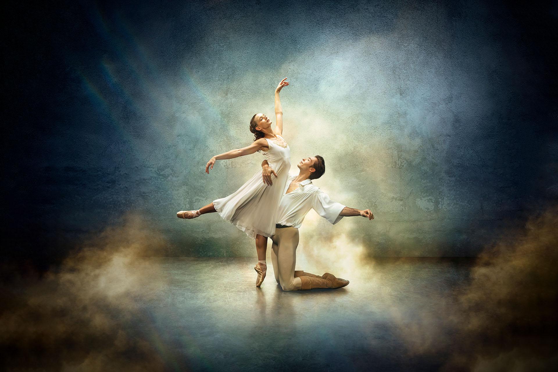 Star-crossed lovers Romeo &amp; Juliet, her standing en pointe and him kneeling in front of her, both wearing white against a fantasy blue and yellow background.