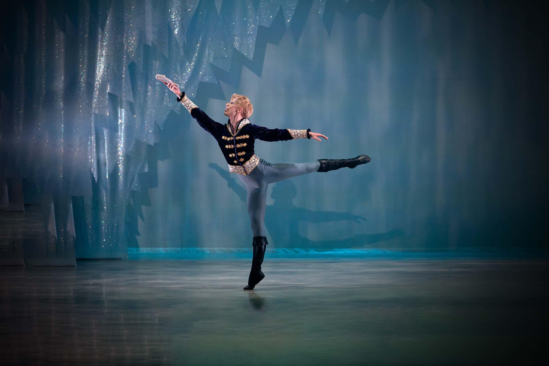 Against an icy blue background, a male dancer stands arabesque holding a silver shoe in front of himself