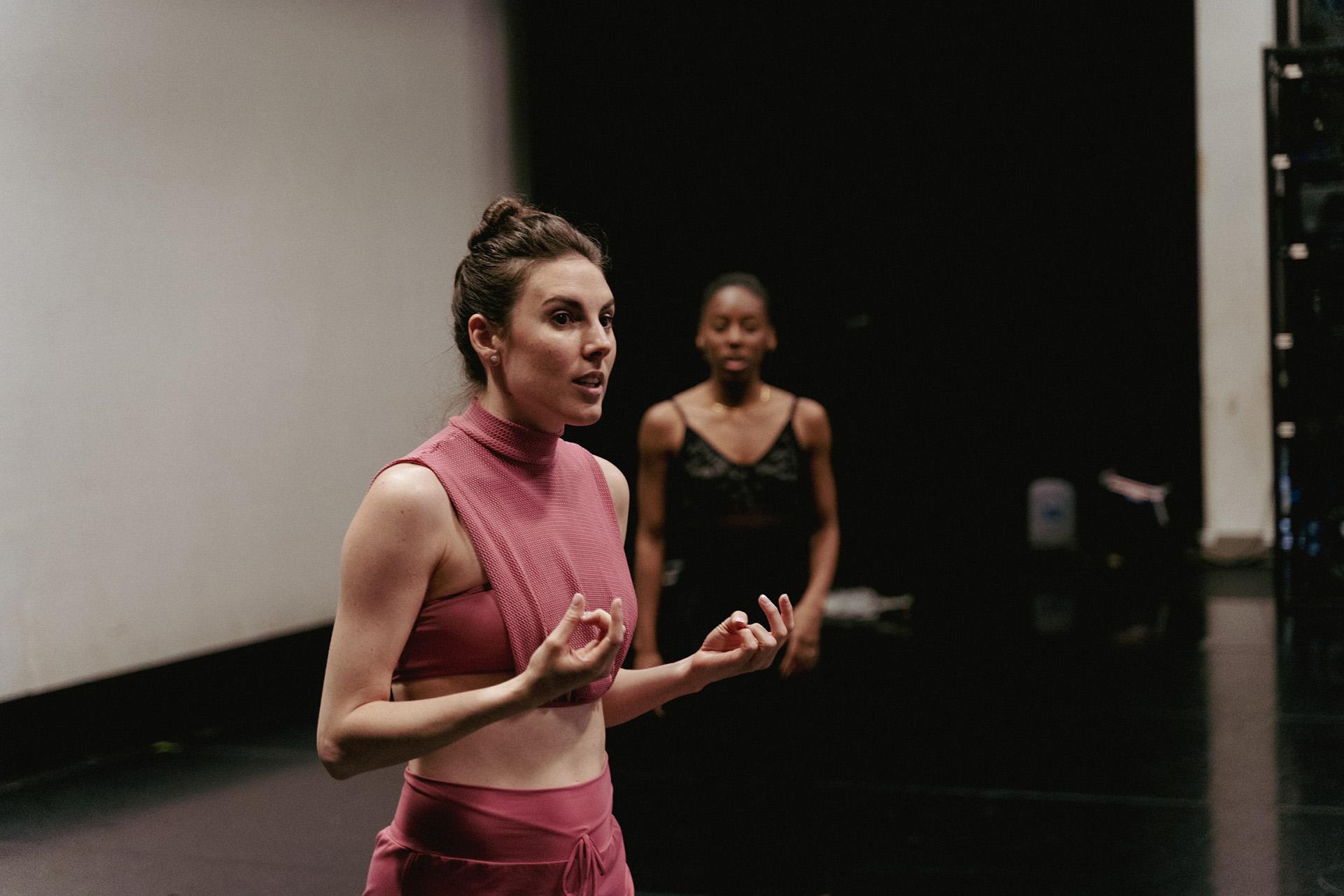 Choreographer wearing a pink top and trousers explaining the movement she envisions to people out of frame