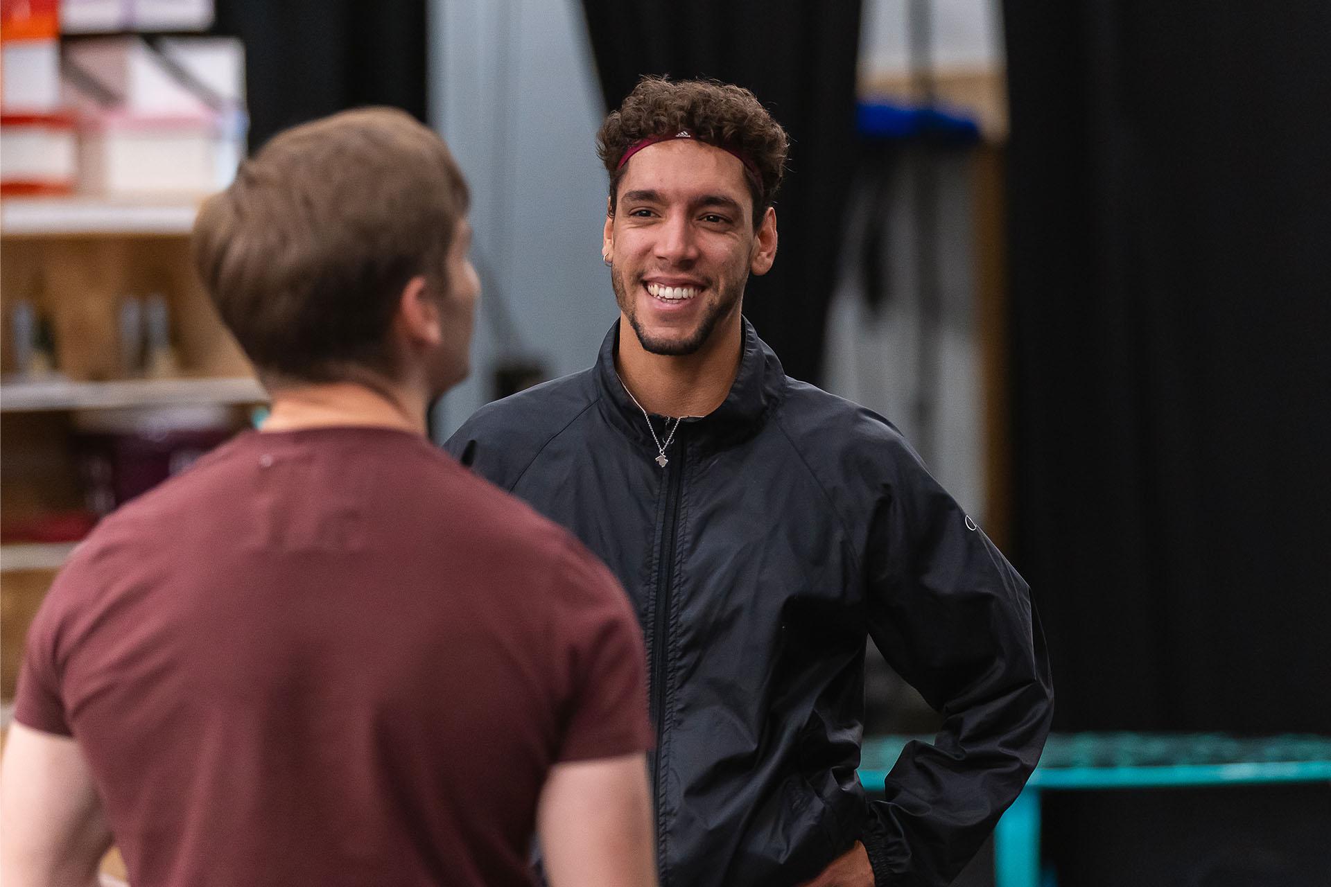 Dancer in a dark zipped up top smiles warmly as he is given instruction and advice