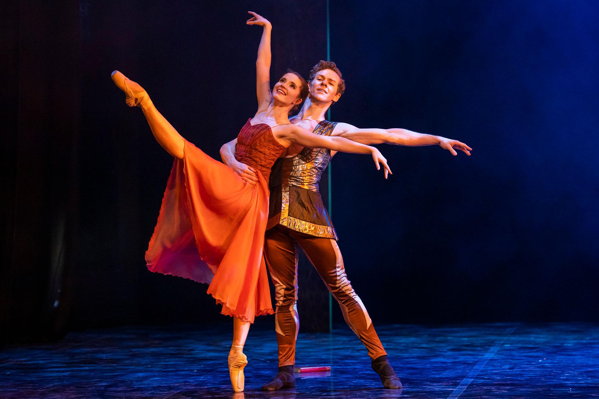two dancers on stage in a ballet pose wearing orange, gold and brown costumes