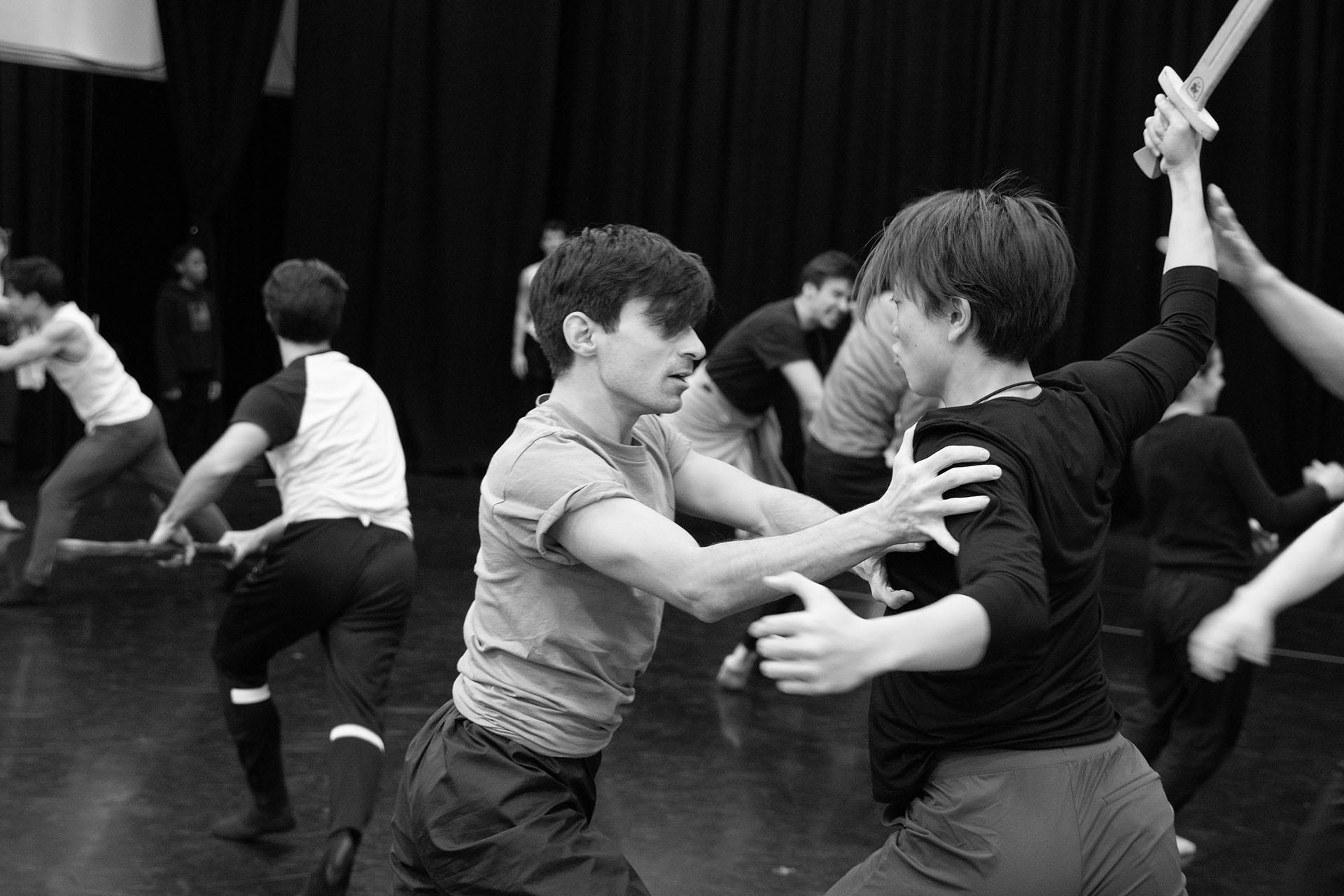 Black and white photo from rehearsals with one man attacking another with a wooden sword while the other holds him at bay