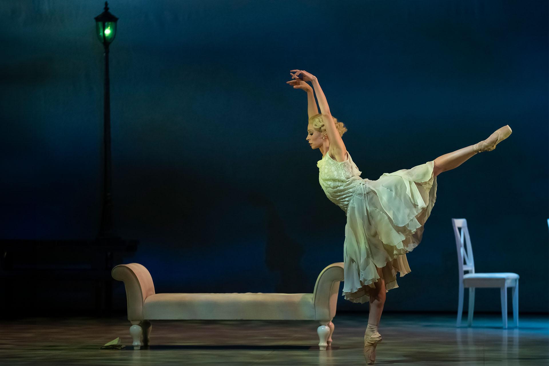 Dancer stands arabesque with her arms above her head wearing a white dress
