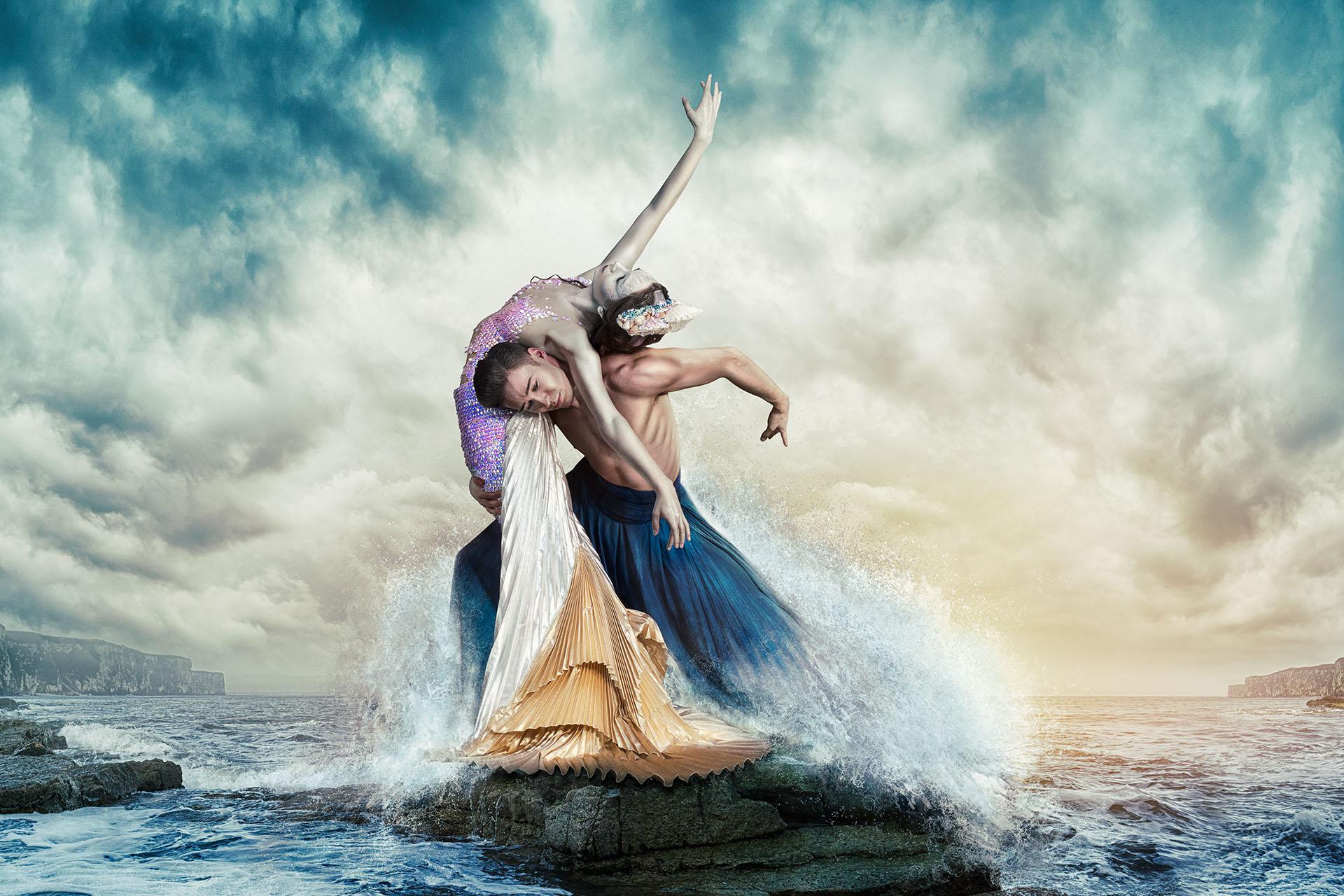 A Mermaid, pale green skin, pink-purple-white-gold outfit being held up by man on his shoulders as she leans back over them, all while being splashed by stylised crashing wavess