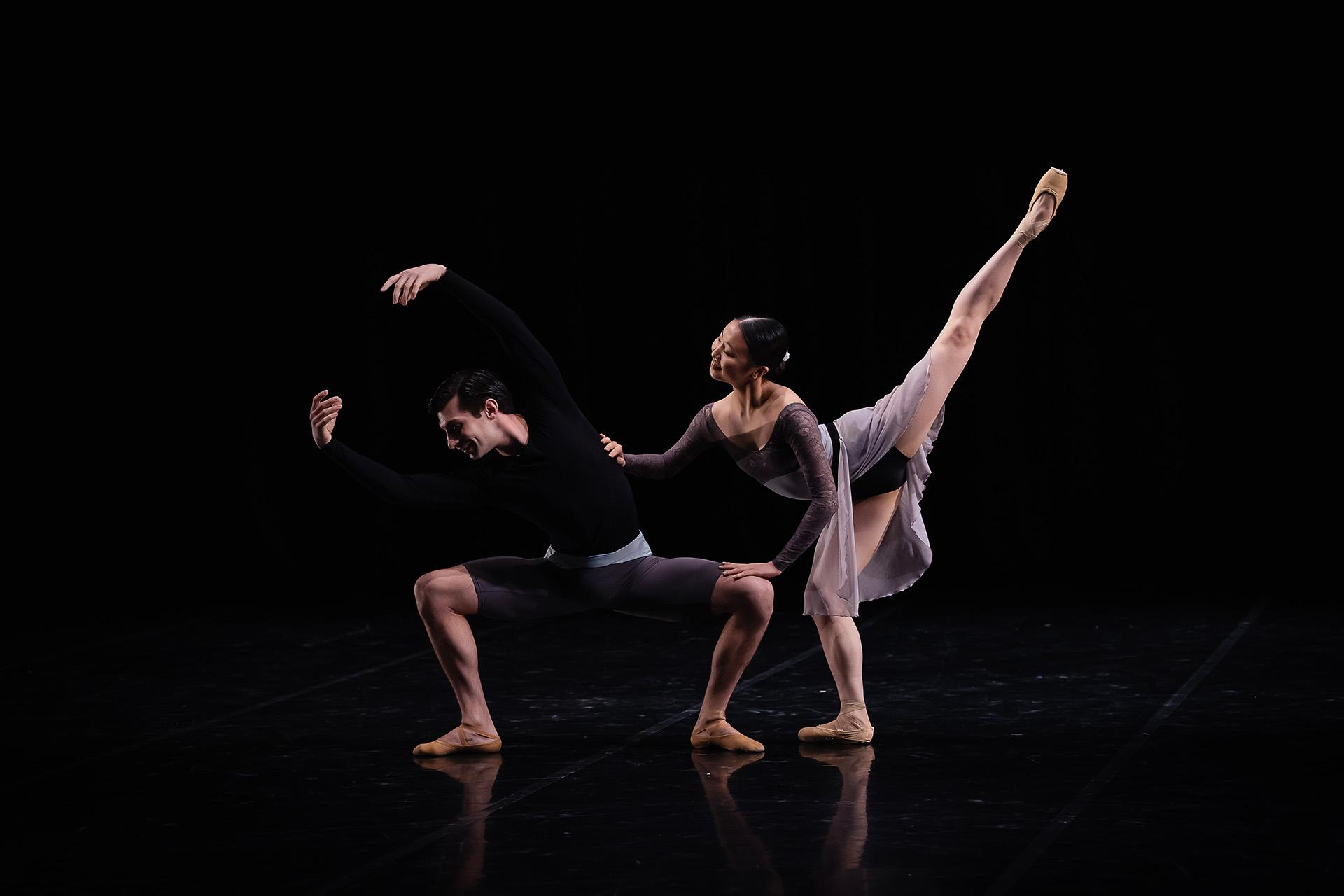 Against a black background two dancer pose, one squatting and arms above his head looks away from the dancer resting a hand on his shoulder with a leg raised straight in the air