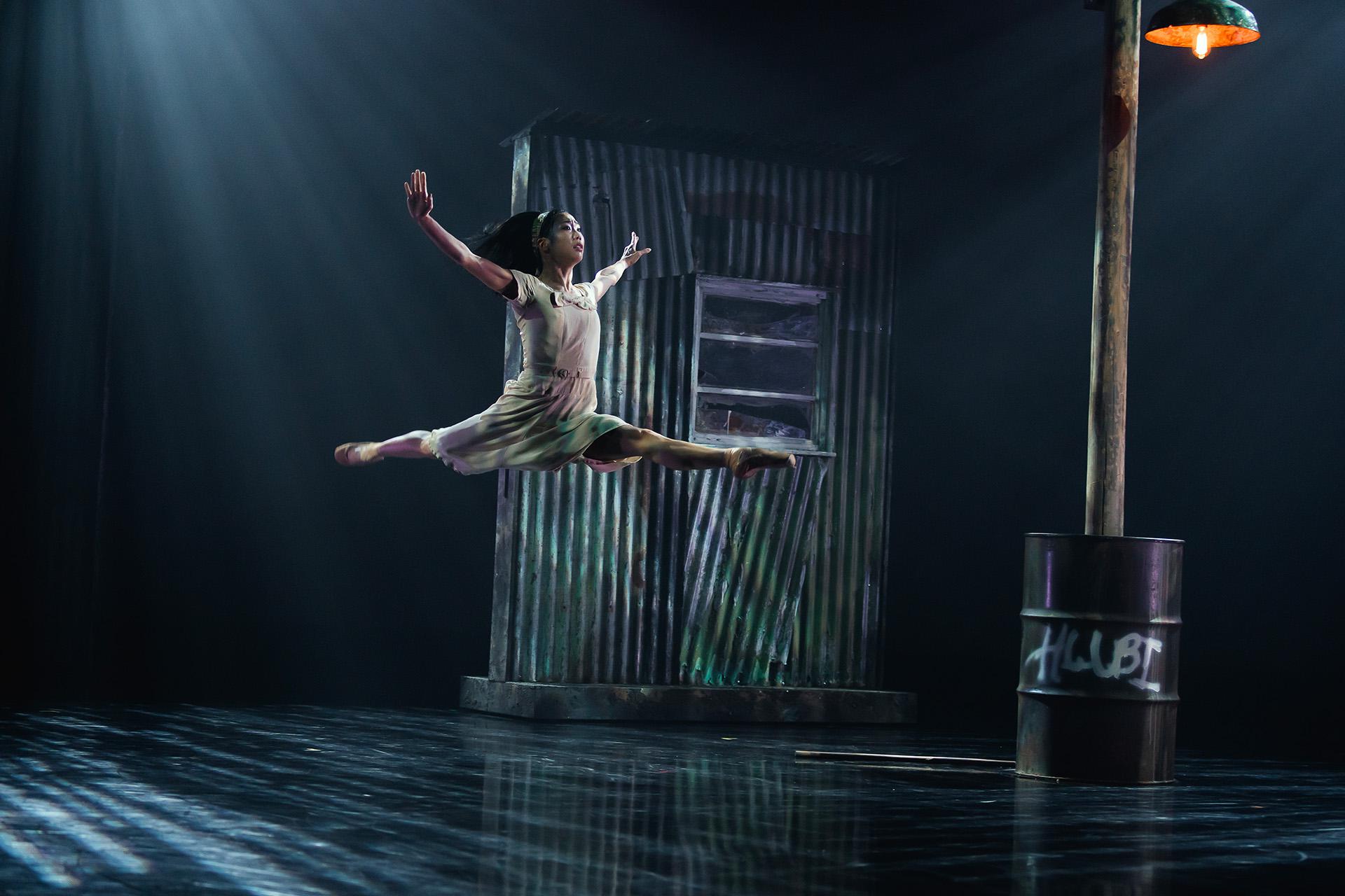 Dancer wearing a light dress leaps gracefully high above the stage, the set of a ramshackle building behind her