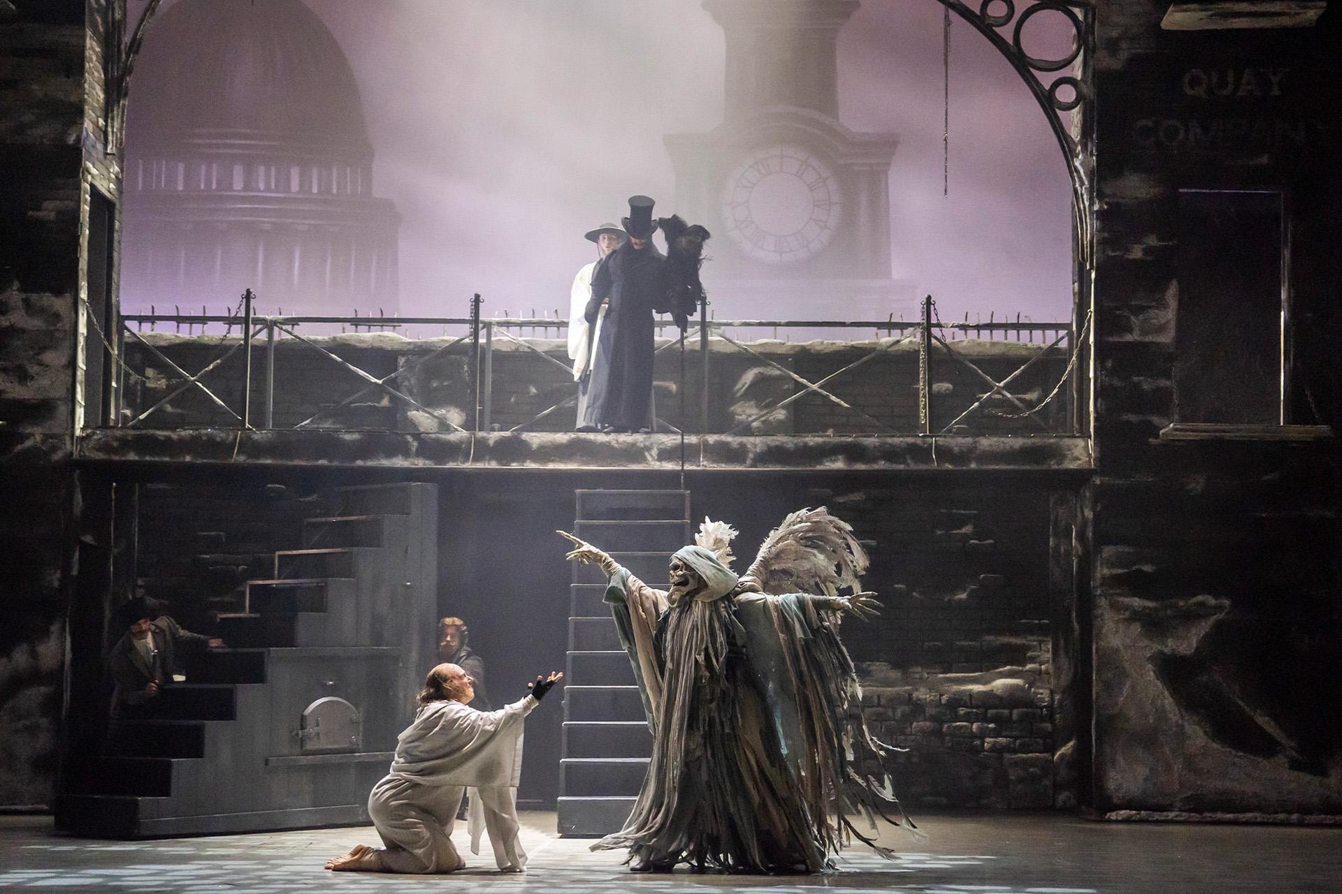 Scrooge begs at the feet of the Ghost of Christmas Yet to Come. A figure dressed in black and a top hat stands at the top of a set of stairs behind them.