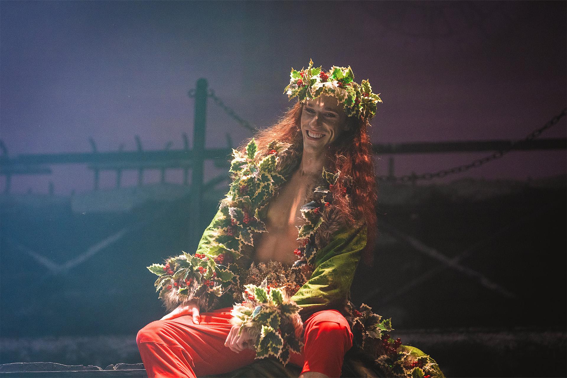 A dancer smiling on stage in a large green and red robe covered in holly with a holly crown
