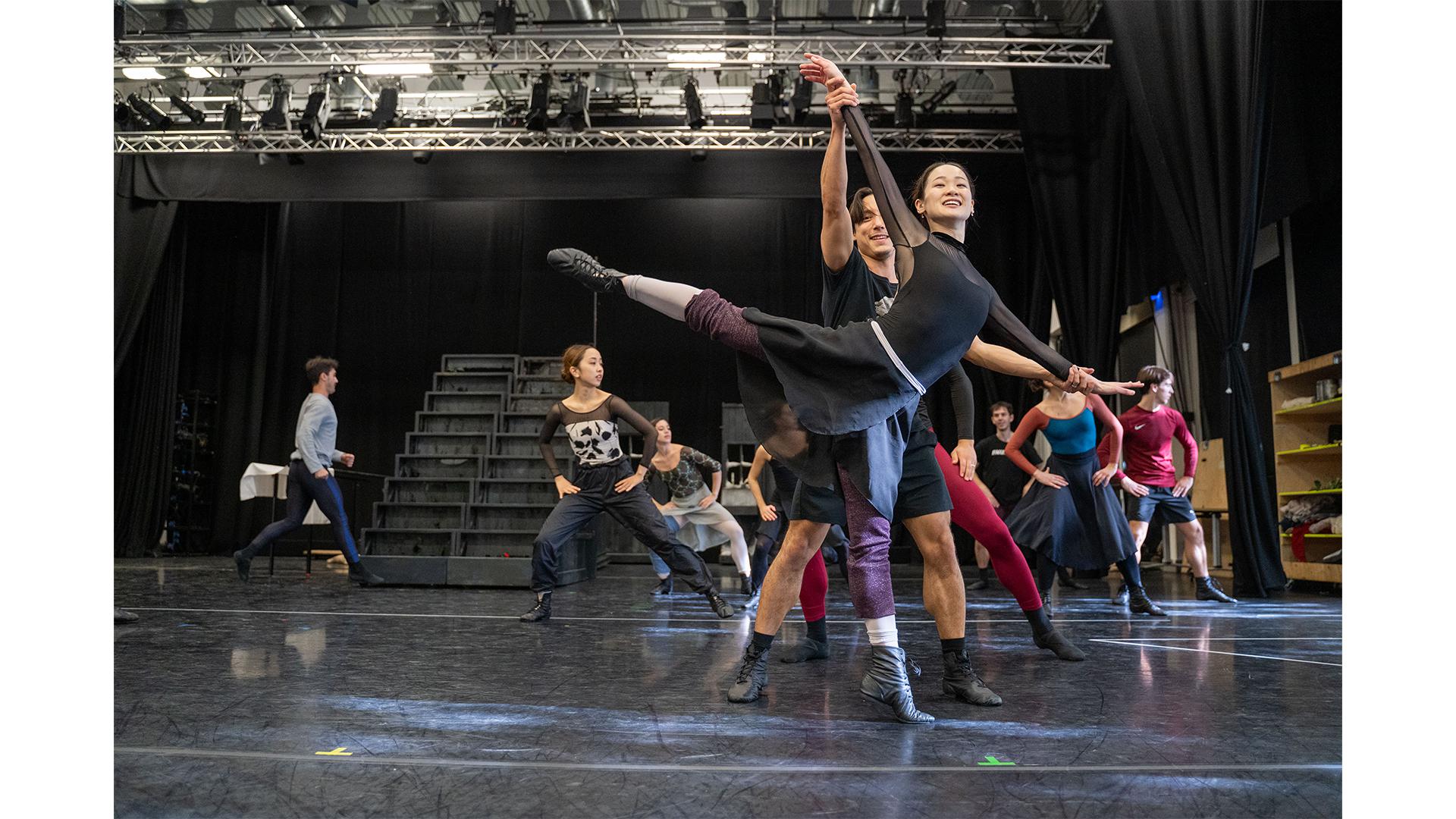 Two dancers stood in the front of frame. One dancer is stood on a single leg while the other supports her.