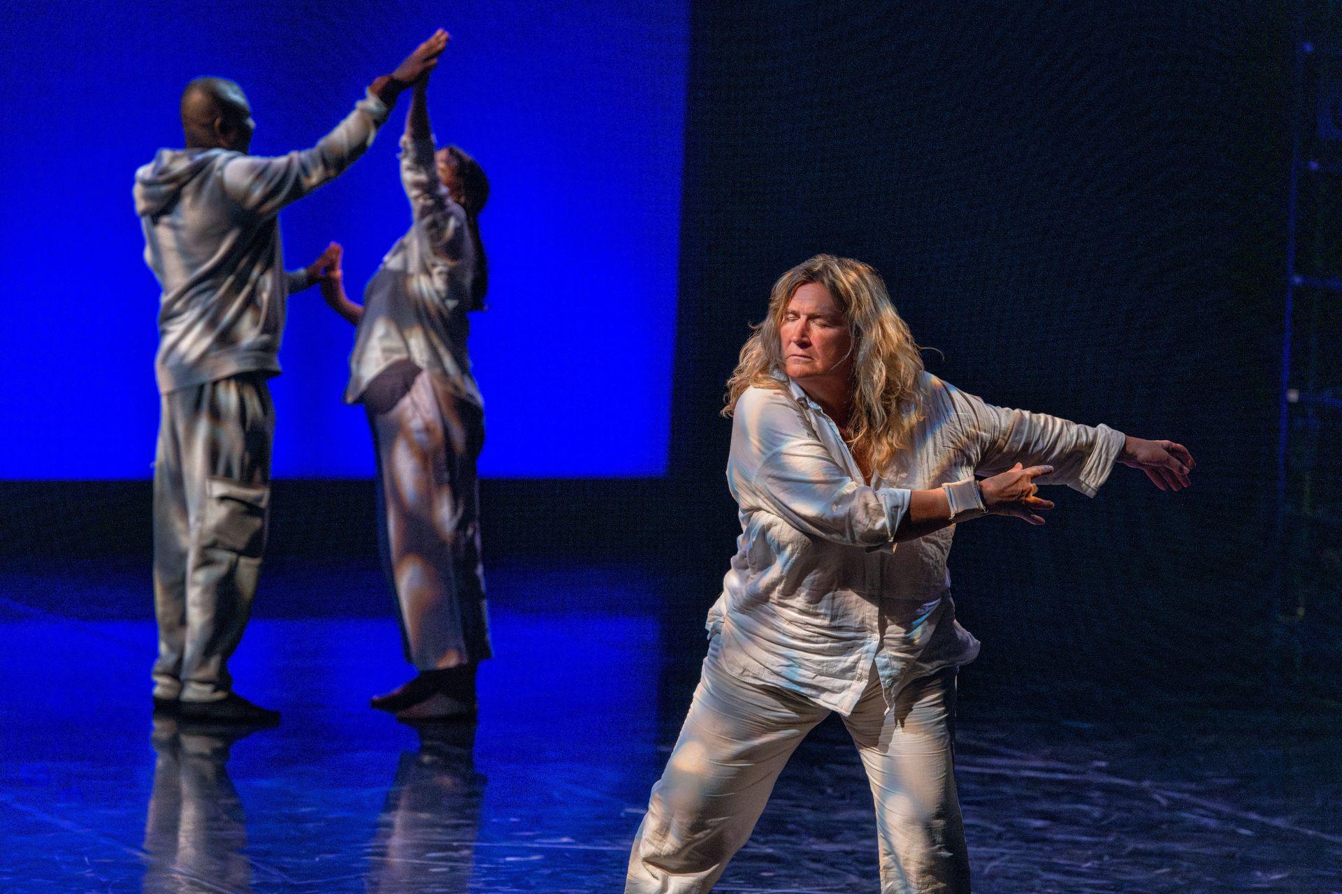 A dancer in pyjamas closes her eyes and sways her arms in front of her. Two other dancers in pyjamas can be seen touching hands behind her.