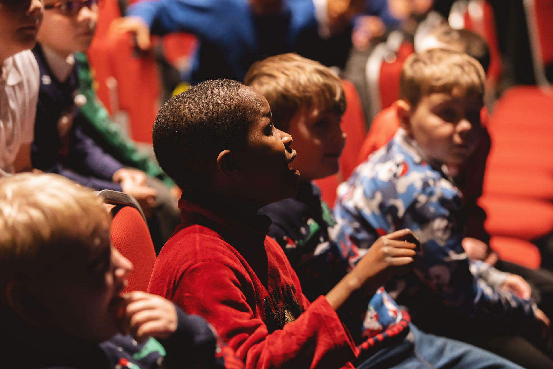 Children in Christmas jumpers sit in an auditorium. One boy opens his mouth to speak to someone out of frame.