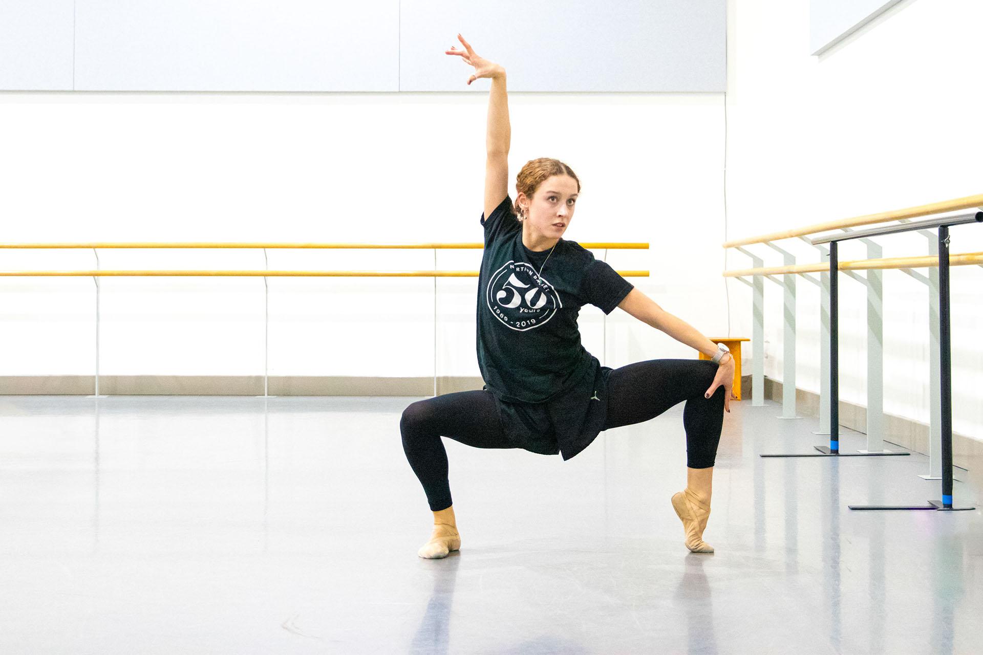 Dancer in rehearsal room crouching, one arm reaching high, the other straight on her knee