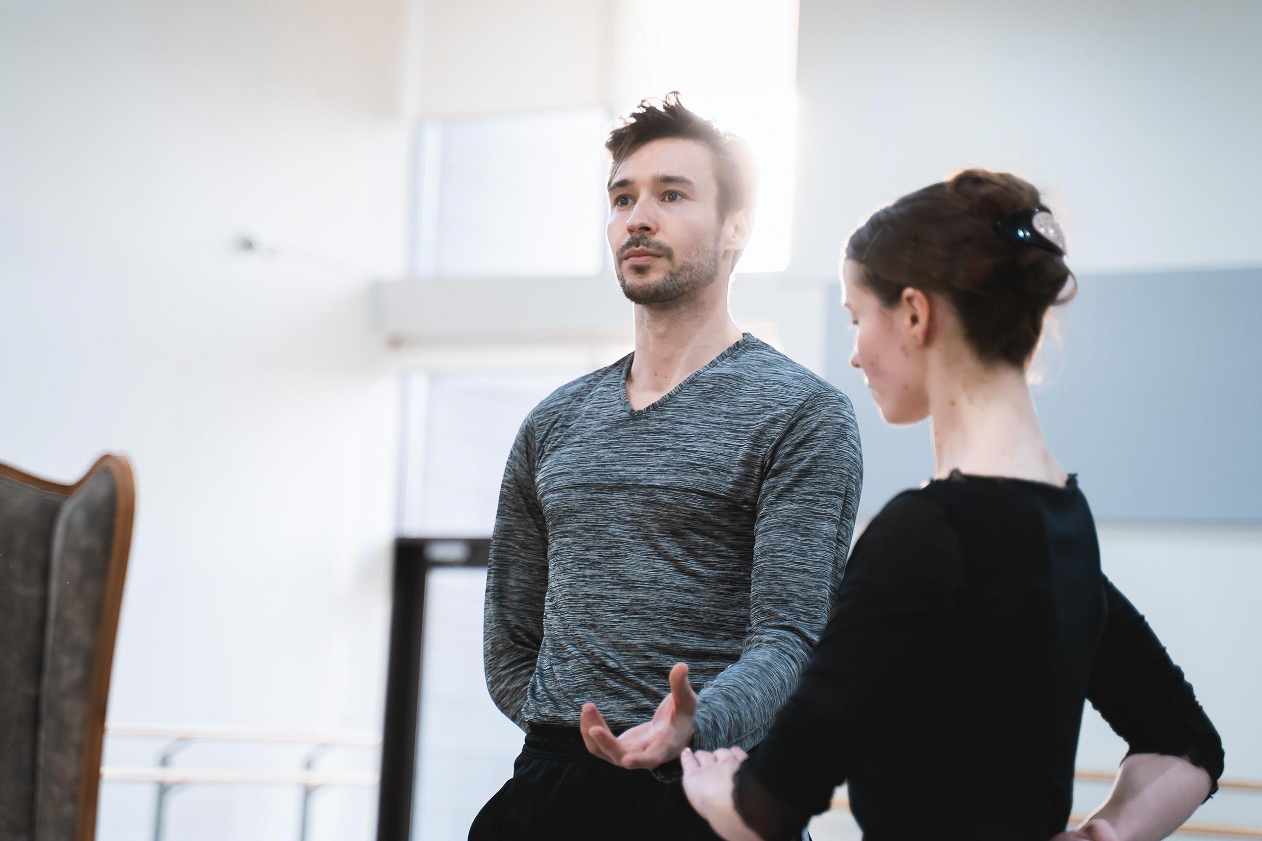 Two dancers in rehearsal, one stands tall - holding a hand out