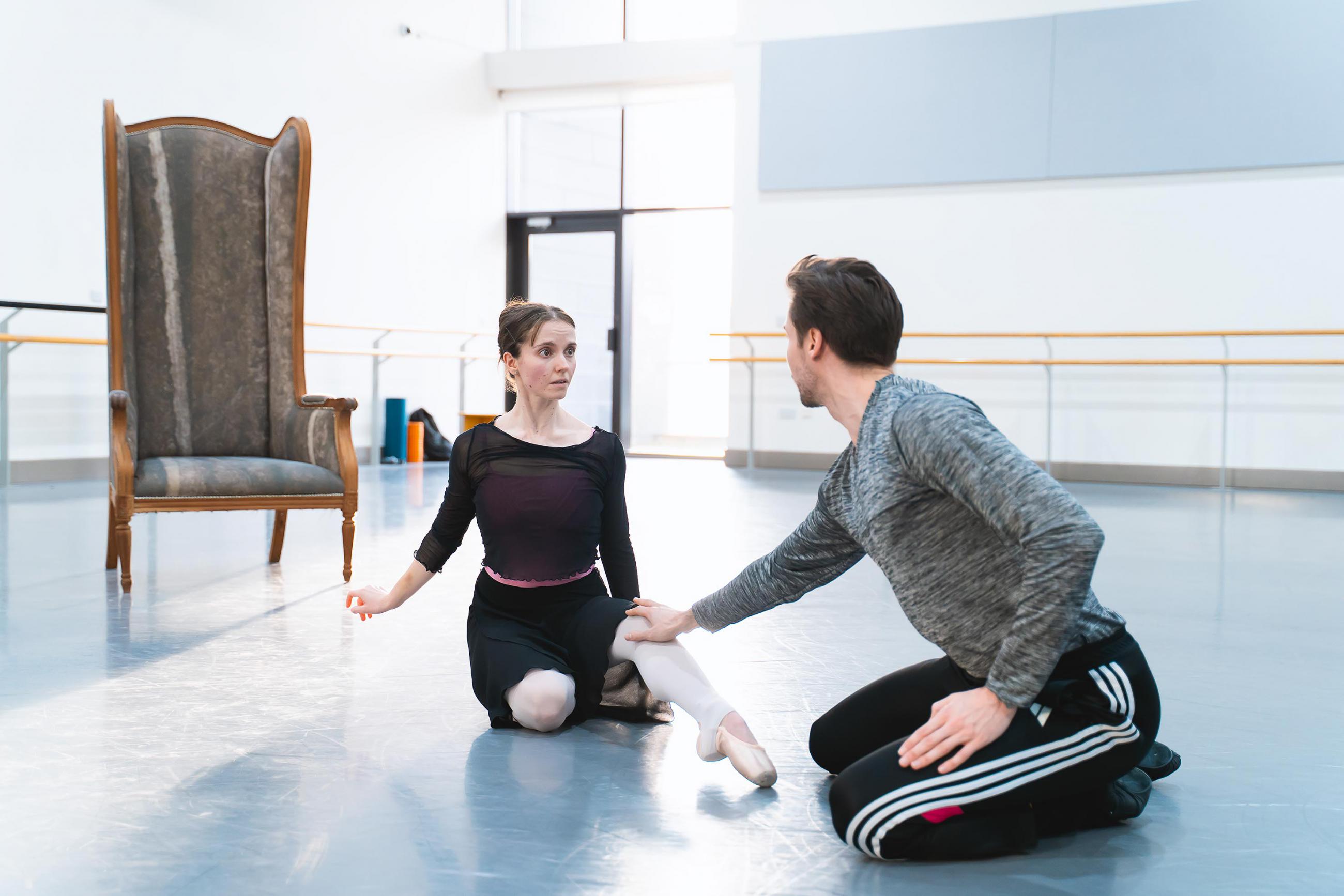 Two dancers rehearsing in a white studio, both sat on the floor. One is reaching towards the other.