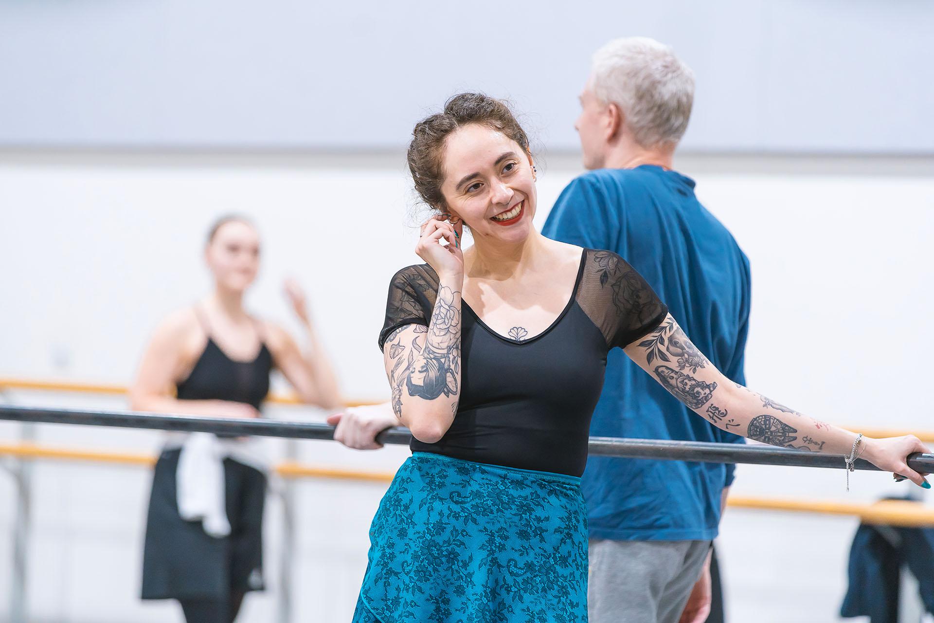 Woman in black top and blue slip stands at the barre, smiling