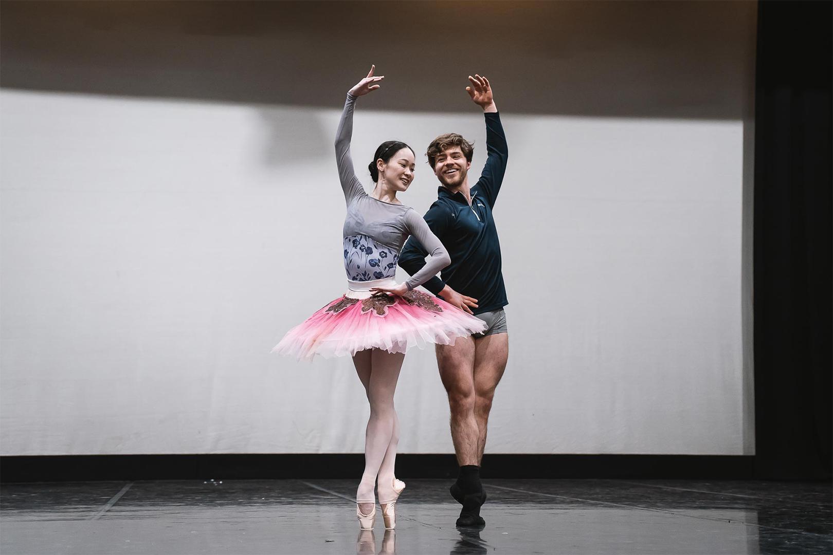 Two dancers stood on their toes with one arm raised looking at each other and smiling