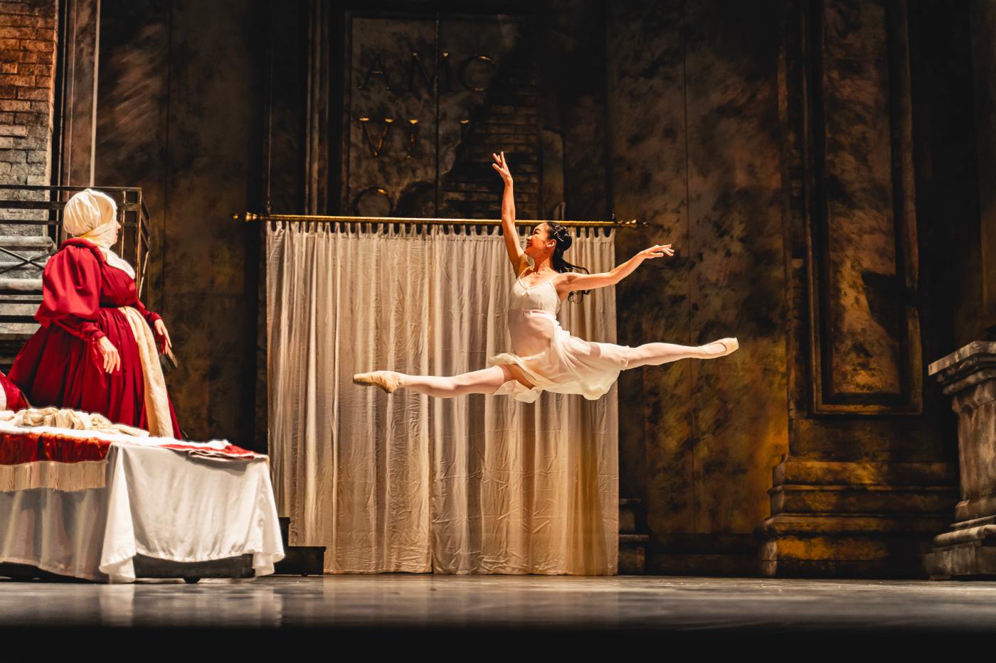 A dancer in a white dress leaps across the stage with her legs in a graceful split position and her arms extended upwards.