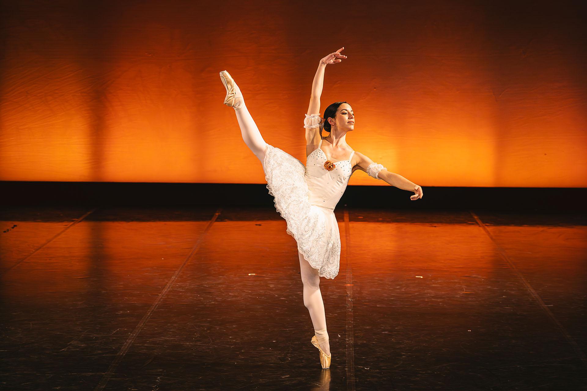 A woman in a white tutu stands battement, one foot en pointe and the other raised straight