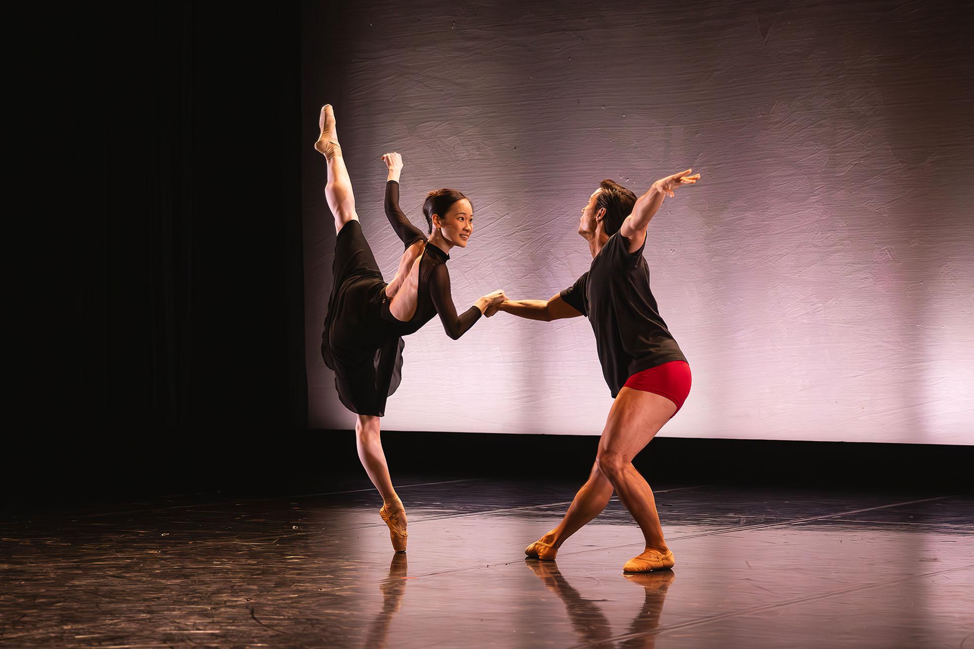 Two dancers of different genders, a female en pointe in a lifted arabesque, a male holding he balance and leaning slightly towards her