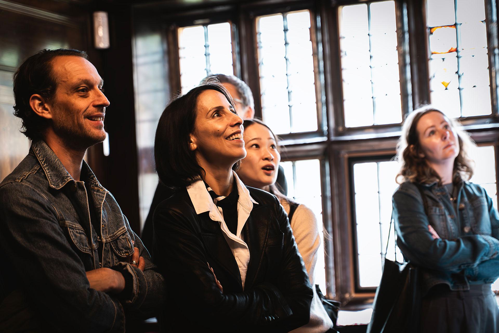Inside Shibden Hall, lit by a large ornate window, Northern Ballet staff, dancers, and Annabelle Lopez Ochoa (Gentleman Jack choreographer) learn about the history of the building and its former denizens