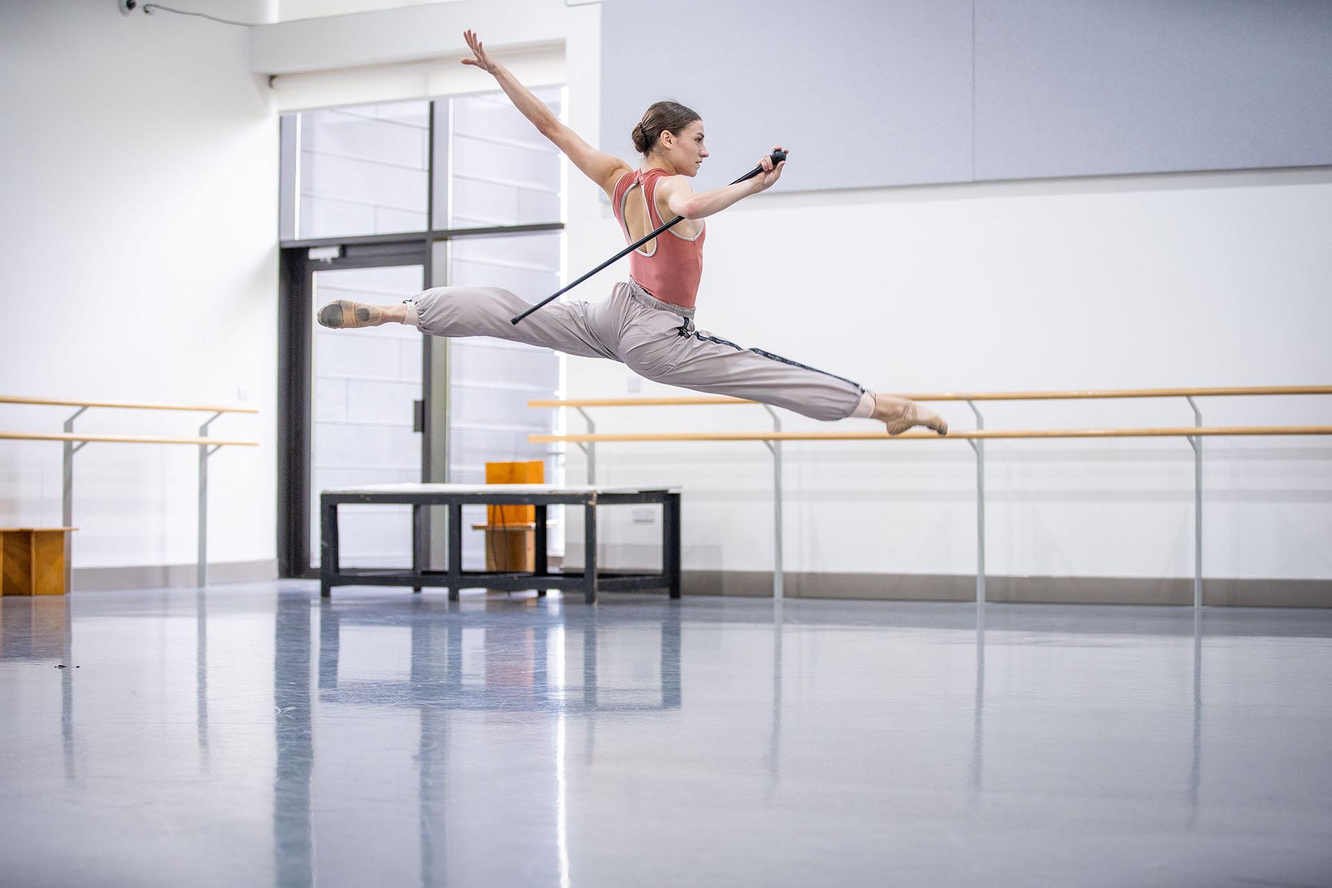 A dancer leaps high, one leg straight in front, the other straight behind, arms above her shoulders and holding her walking stick in one hand