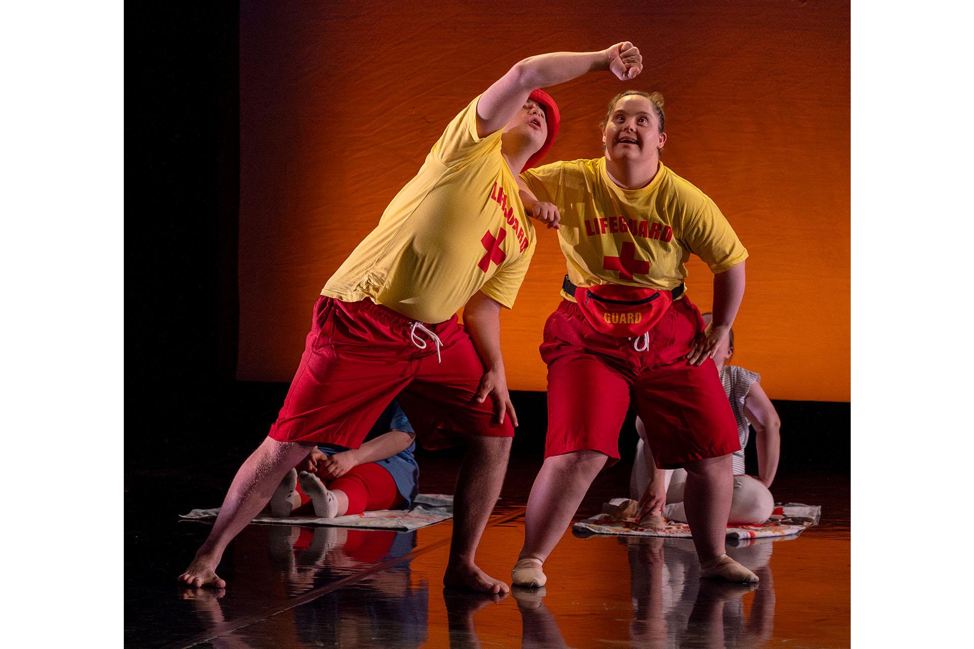 Two dancers from the Ability Class stand on stage wearing life guard uniforms of yellow t-shirt and red shorts