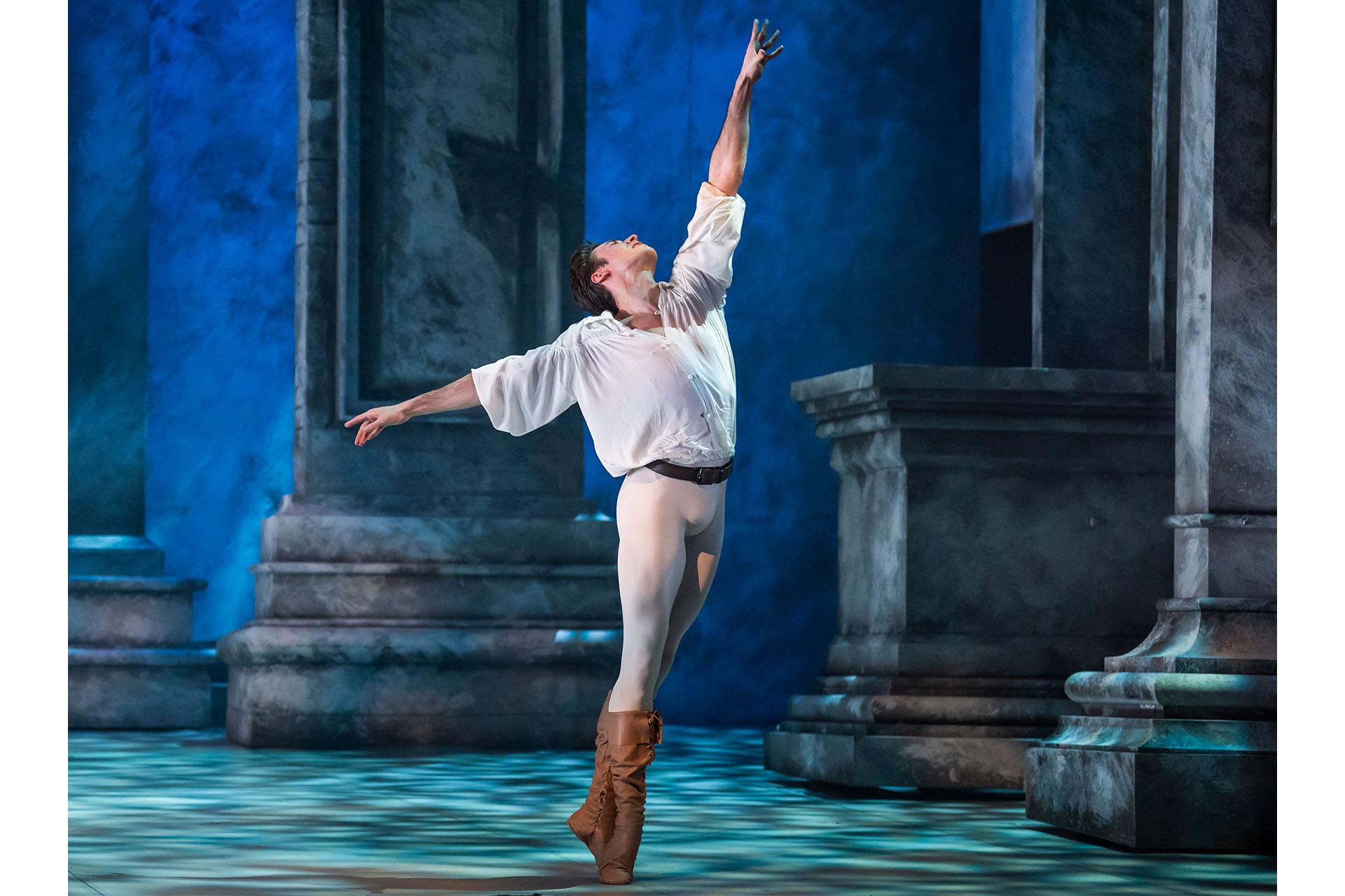 A man in leather boots, light trousers, dark belt and white shirt stands forlorn, one hand raised high, the other stretched behind his, his head thrown back