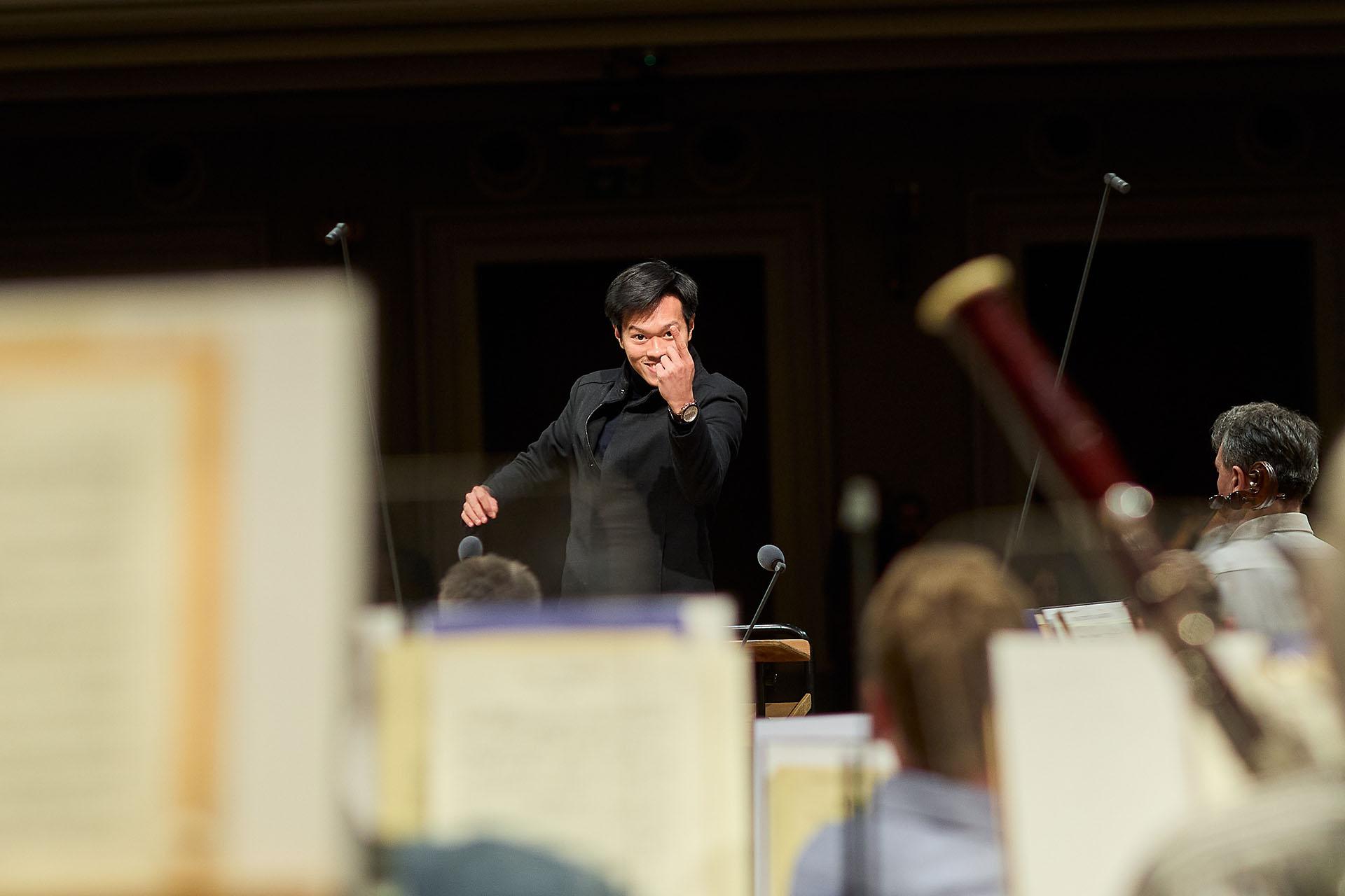 Conductor with a raised hand wearing black, viewed over musicians