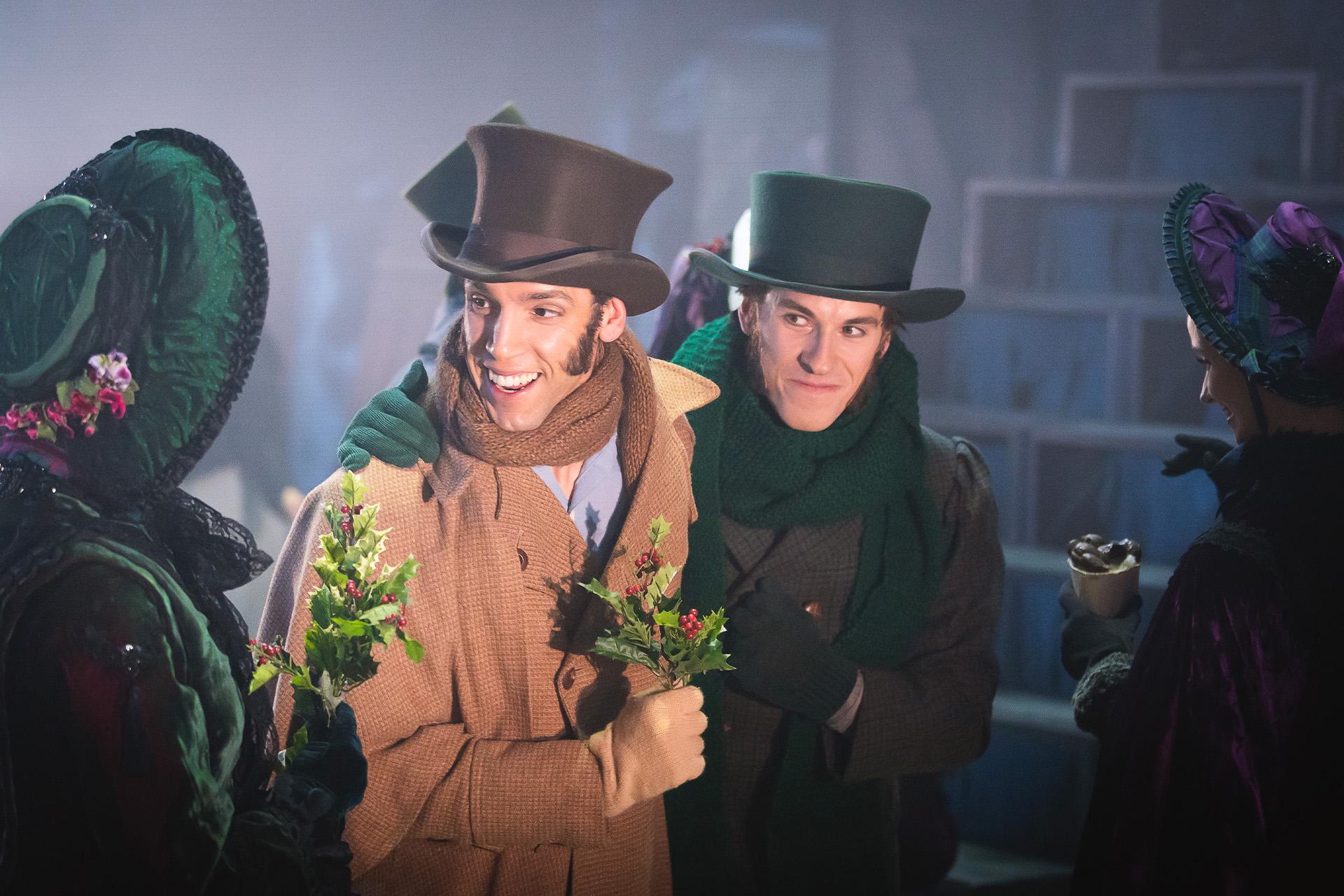Two dancers in Dickensian costumes interacting on stage, both holding a leaf of holly