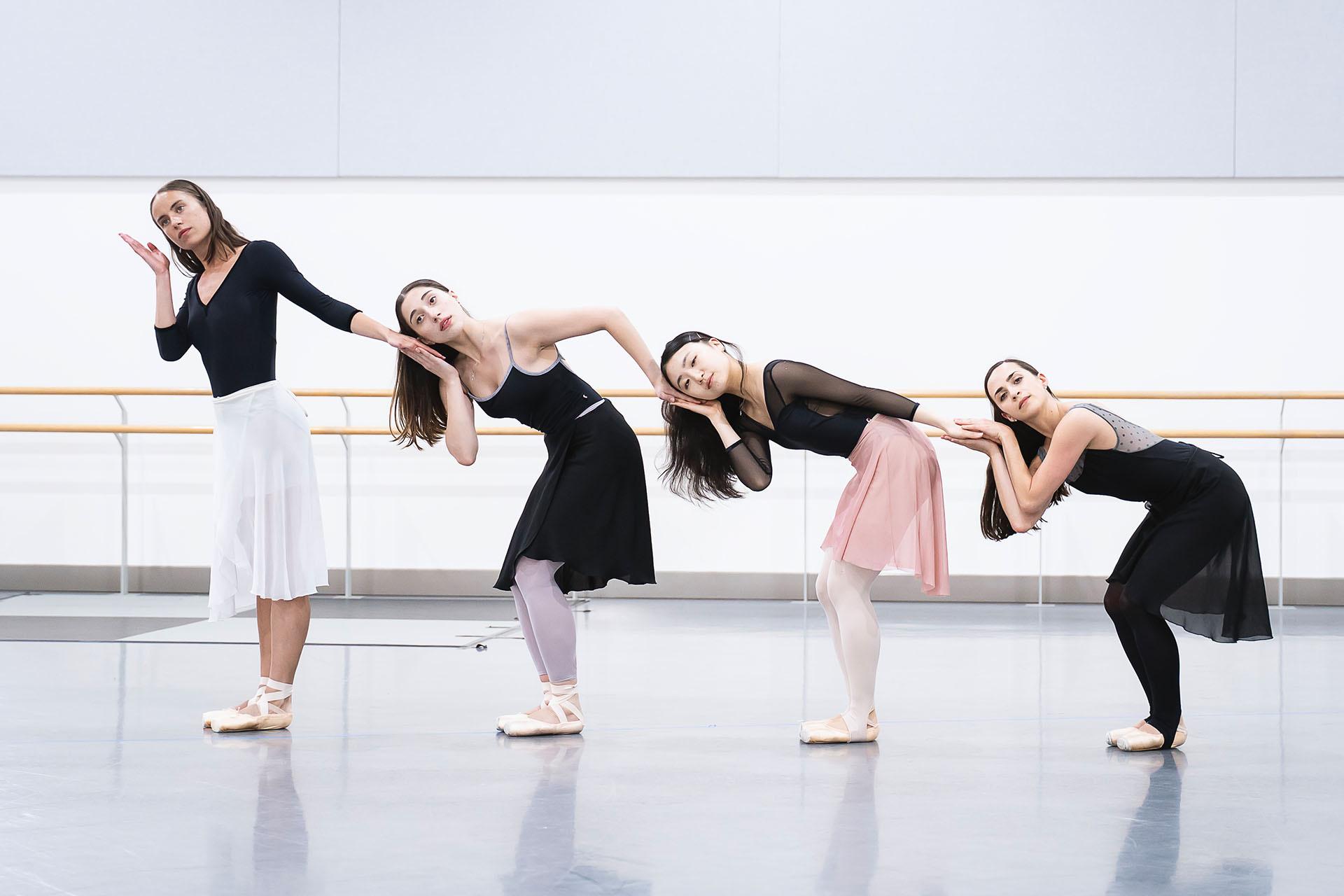 Four dancers in a row, half-turned towards the camera, each leaning lower than the woman in front of her and each holding hands with the person behind
