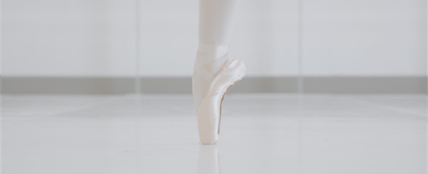 A single foot stands en pointe on a white studio floor.
