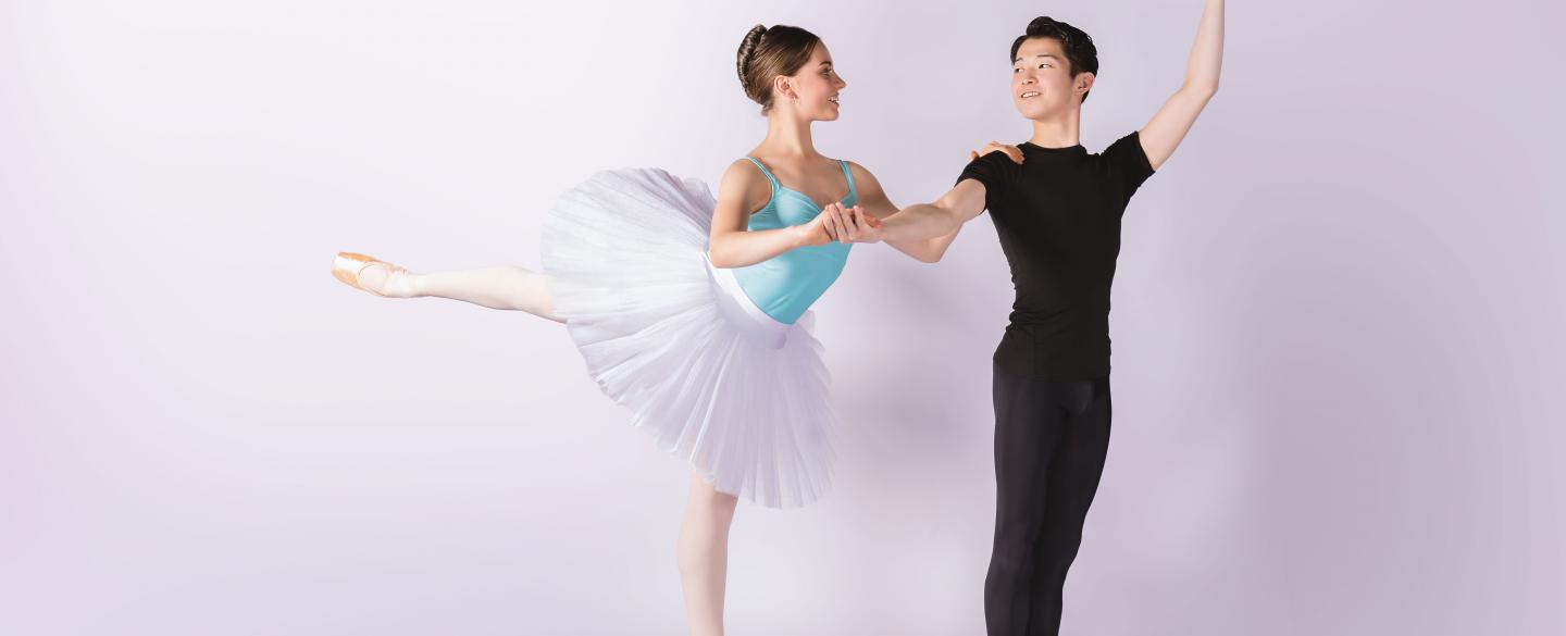 A female student in a white tutu stood on pointe holding the arm of a male student dressed in black