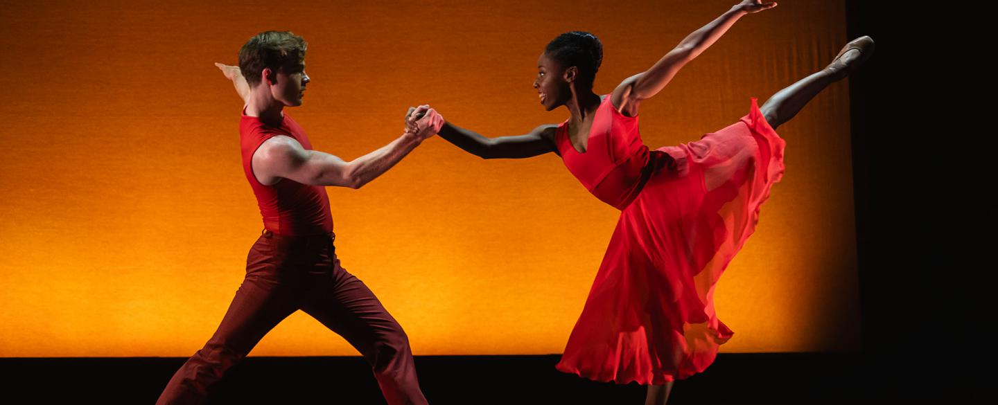 Woman balances en pointe on one foot, leg behind her raised high and another dancer helping her balance by holding her right hand