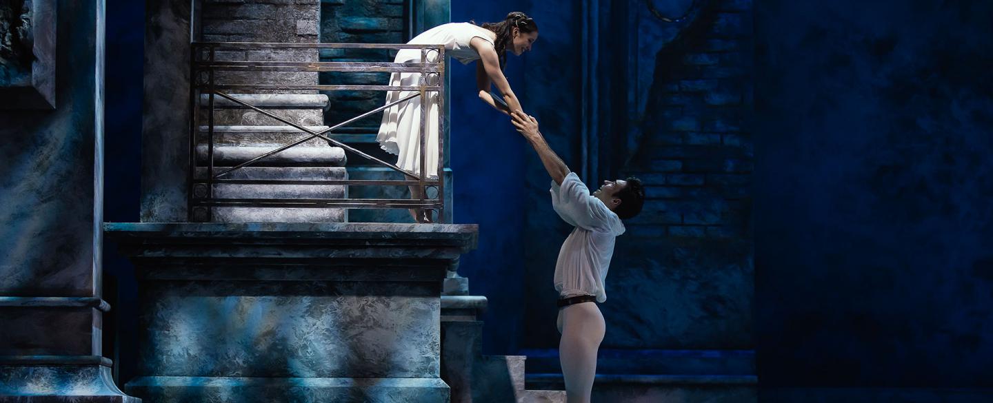 Juliet leans over her balcony and Romeo reaches up to hold her hand